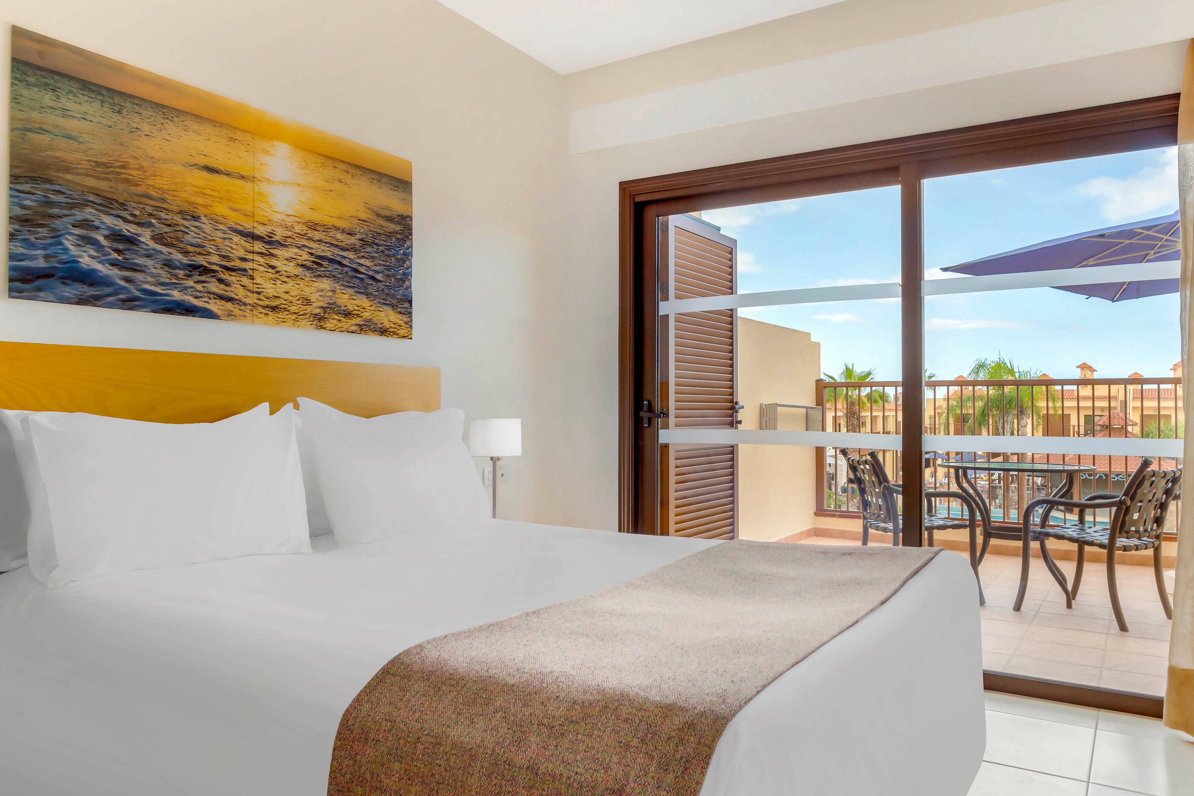A neatly made hotel bed with white linens dominates the foreground of a bright guest room. A large sliding glass door opens onto a sunny terrace with outdoor chairs and a table. Beyond the terrace, palm trees and warm-toned buildings suggest a resort setting. Soft natural light and a seascape wall art create a calm, inviting atmosphere ideal for relaxation.