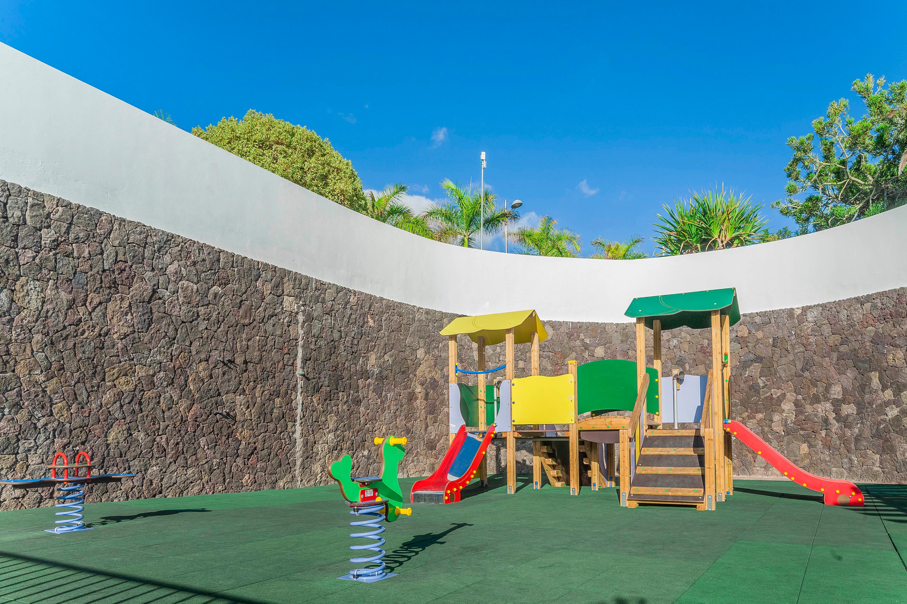 A small outdoor playground with colorful play structures stands inside a high stone-walled courtyard. The equipment includes slides, climbing platforms, and spring riders on a green safety surface. Bright red, yellow, green, and blue elements contrast with the dark stone wall. Palm trees and clear blue sky rise above the curved wall, creating a sunny and inviting atmosphere.