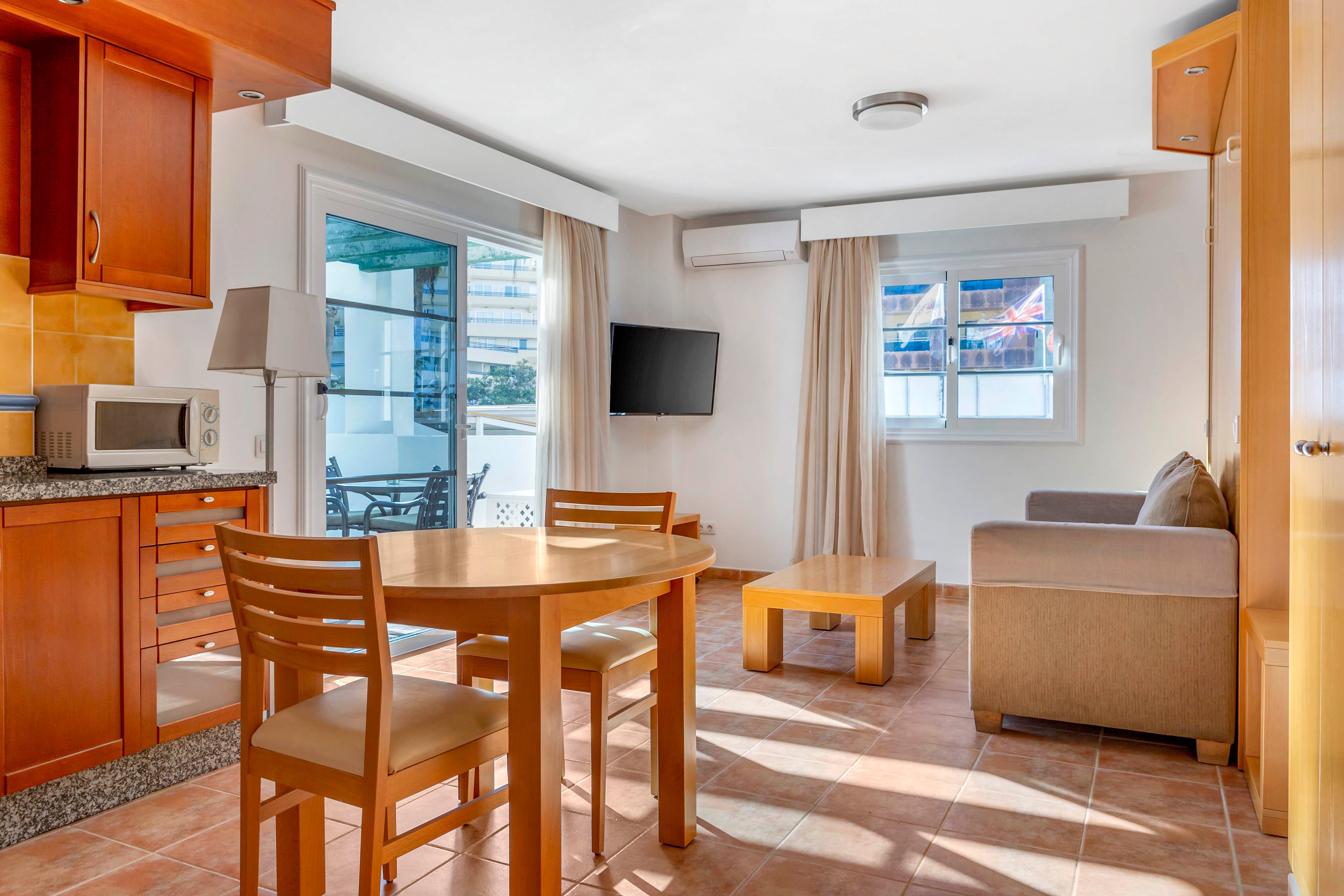 A bright open-plan apartment living area combines a small kitchen, dining space, and lounge. Wooden cabinets and a round dining table with chairs sit beside a sliding glass door leading to a balcony. A sofa and low coffee table face a wall-mounted flat-screen TV, with a window bringing in additional daylight. Neutral walls, tiled flooring, and light curtains create a clean, modern vacation rental feel.