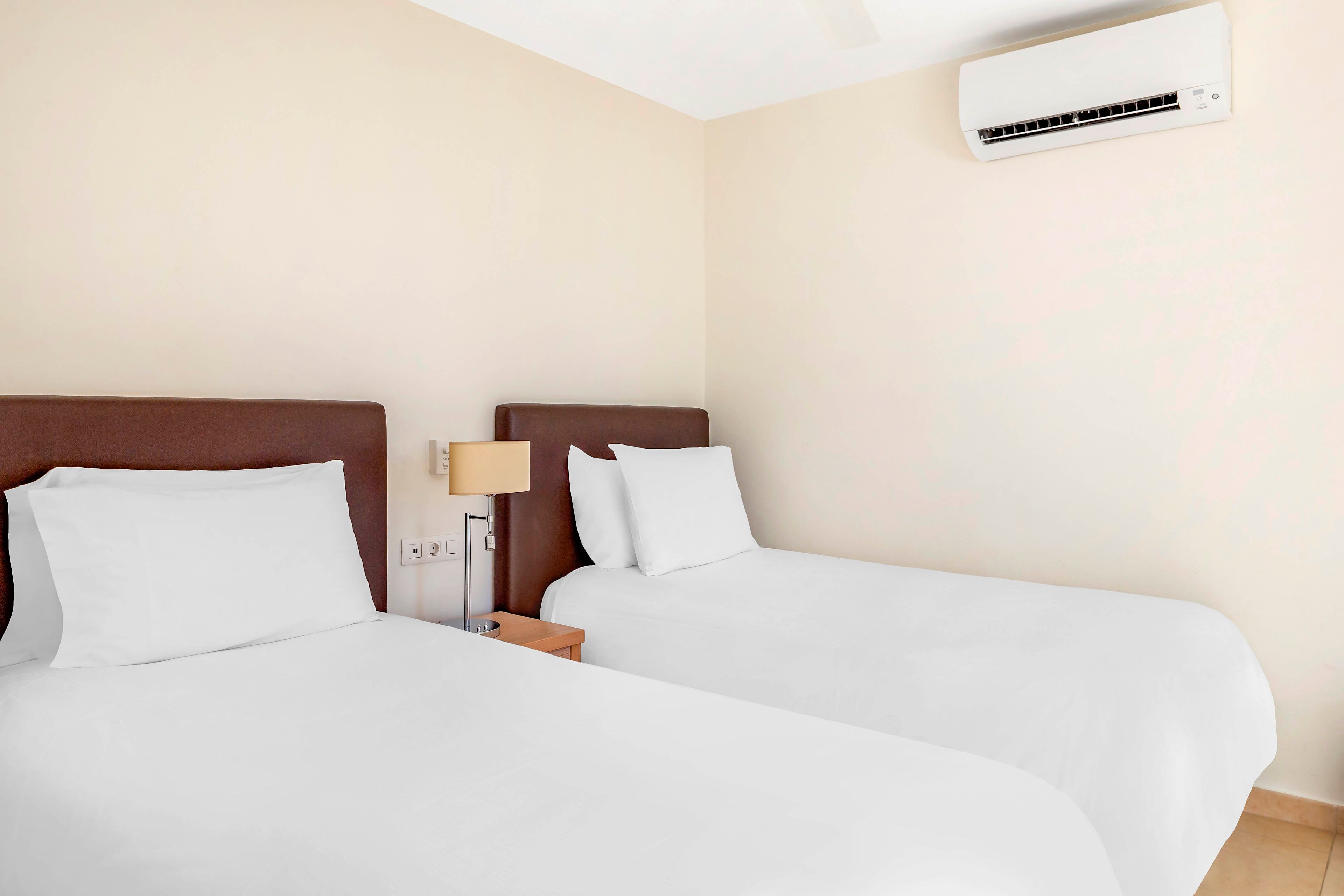 A clean, minimalist hotel room features two neatly made twin beds with white linens and dark headboards. A small wooden nightstand with a lamp sits between the beds against a light-colored wall. An air conditioning unit is mounted high on the wall above the beds, emphasizing comfort and climate control. The overall scene feels bright, tidy, and inviting, suitable for travelers or guests.