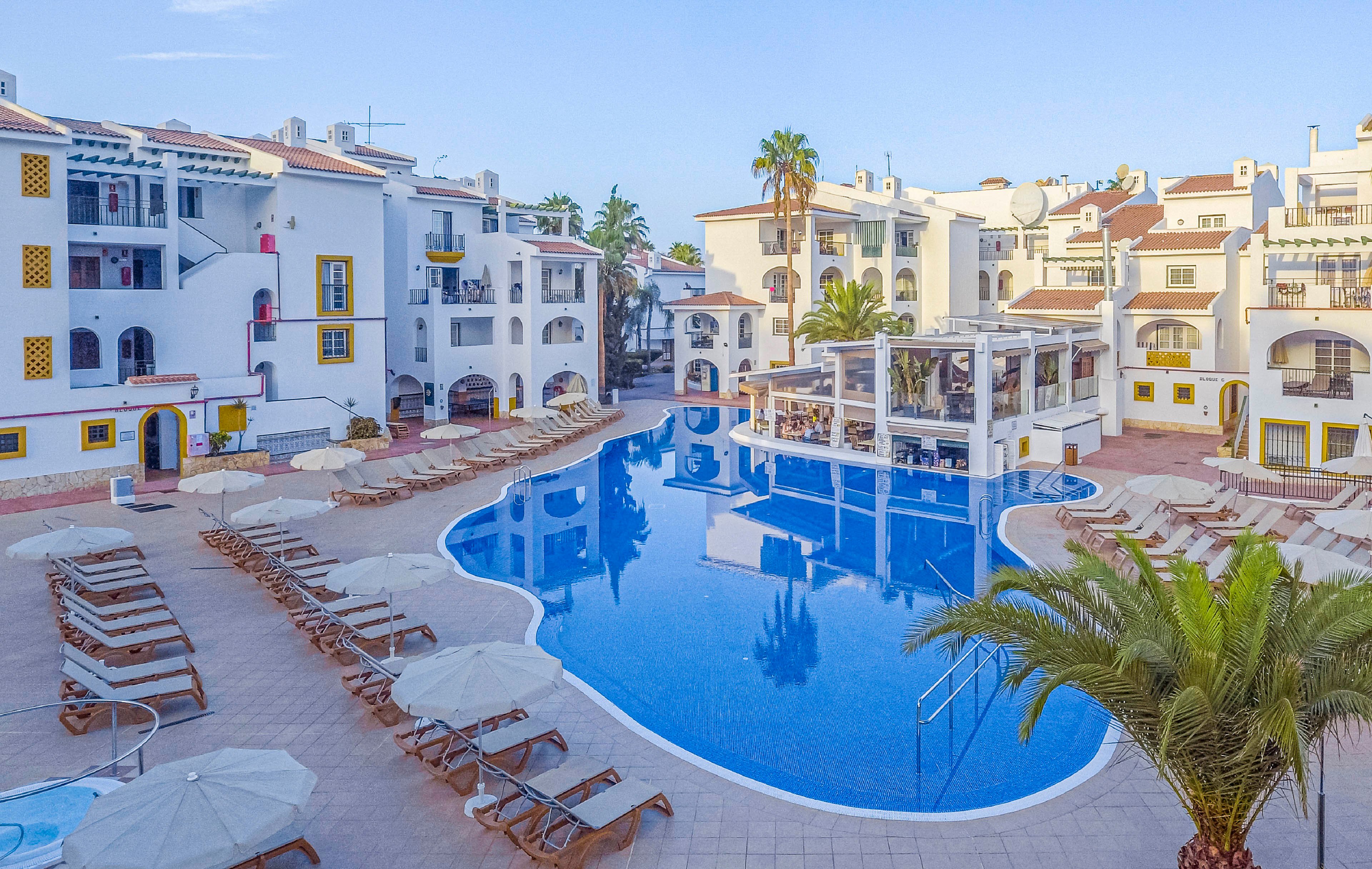 A bright resort courtyard features a large, curved blue swimming pool surrounded by white apartment-style buildings. Numerous sun loungers and umbrellas line the tiled pool deck, suggesting a relaxing holiday setting. Palm trees and balconies add a Mediterranean or coastal atmosphere to the scene. The clear sky and calm water create a peaceful, inviting vacation mood.