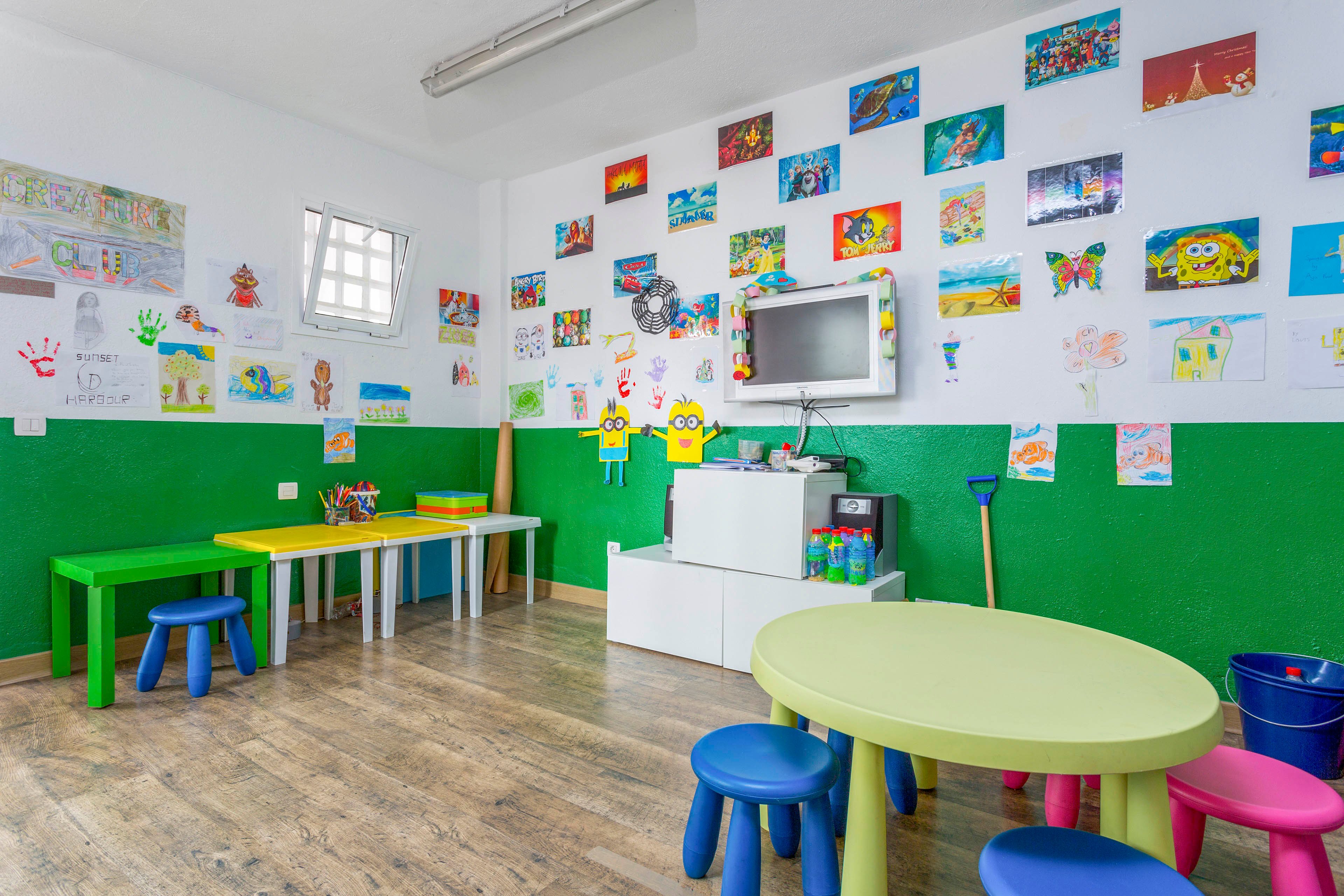 A bright children's playroom features small colorful tables and stools arranged on a wooden floor. The walls are painted white and green and are covered with numerous children's drawings and paintings. A television is mounted on the wall above white storage cubes holding toys and supplies. The room is well-lit and designed to be inviting and creative for young children.