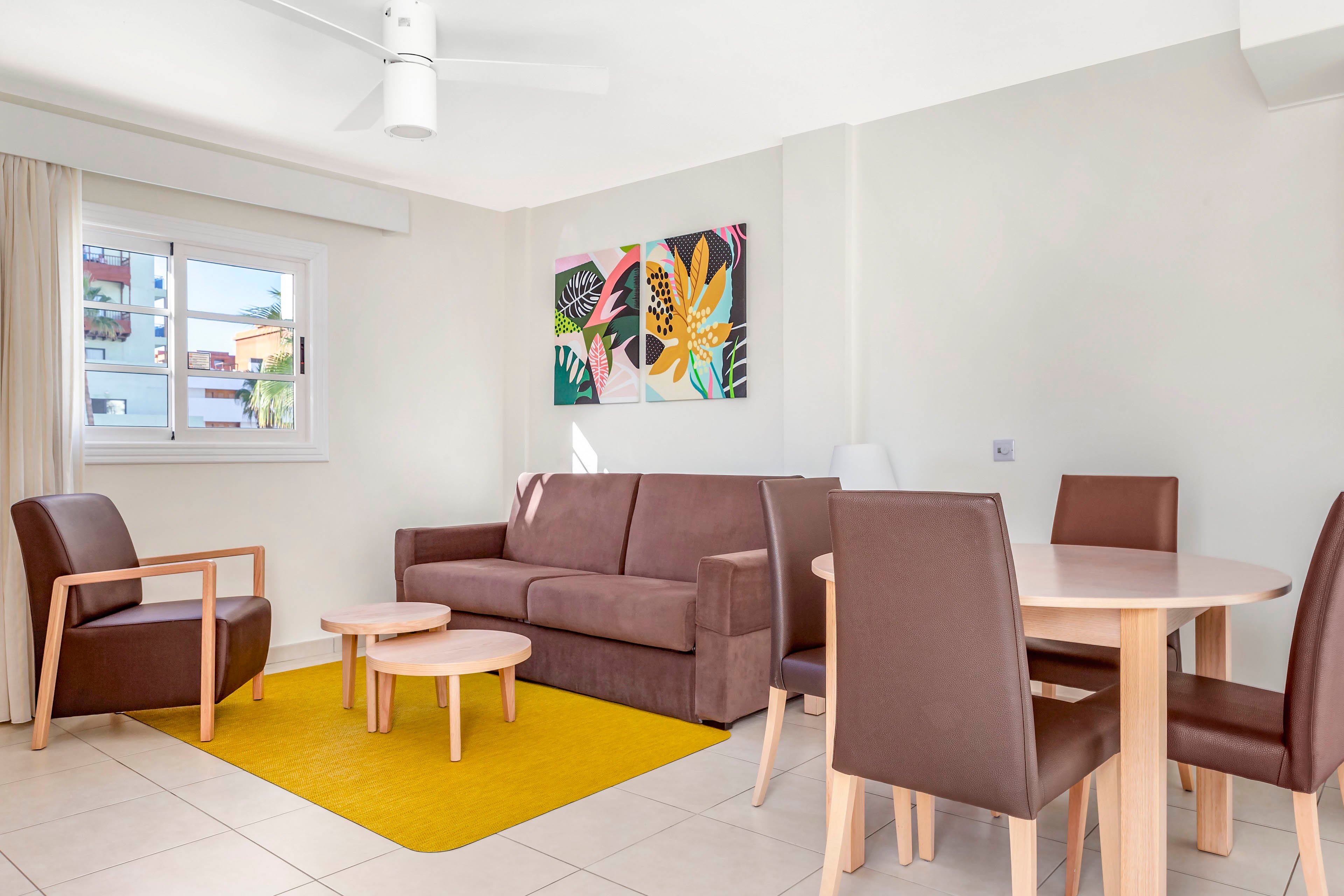 A contemporary living room features a brown sofa and matching armchairs arranged around small nesting coffee tables. A round wooden dining table with four brown chairs sits to the right, creating a compact dining area. Colorful abstract artwork hangs above the sofa, adding vibrant accents to the neutral white walls. Natural light enters through a window with sheer curtains, illuminating the yellow rug and tiled floor.