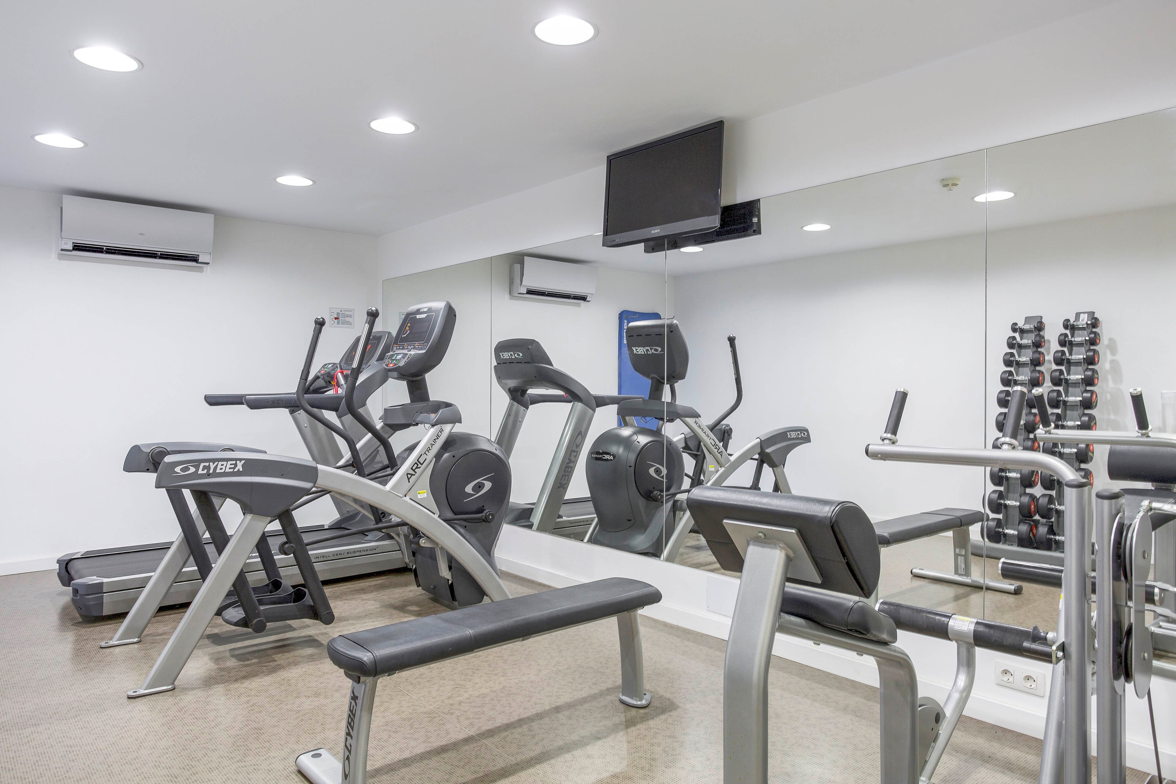 A clean, modern gym room features several exercise machines including treadmills, ellipticals, and weight benches. The space has bright white walls, recessed ceiling lights, and a large wall mirror reflecting the equipment. A rack of dumbbells stands against the mirrored wall, and a flat-screen television is mounted above. The room appears empty, emphasizing the organized layout and contemporary fitness environment.