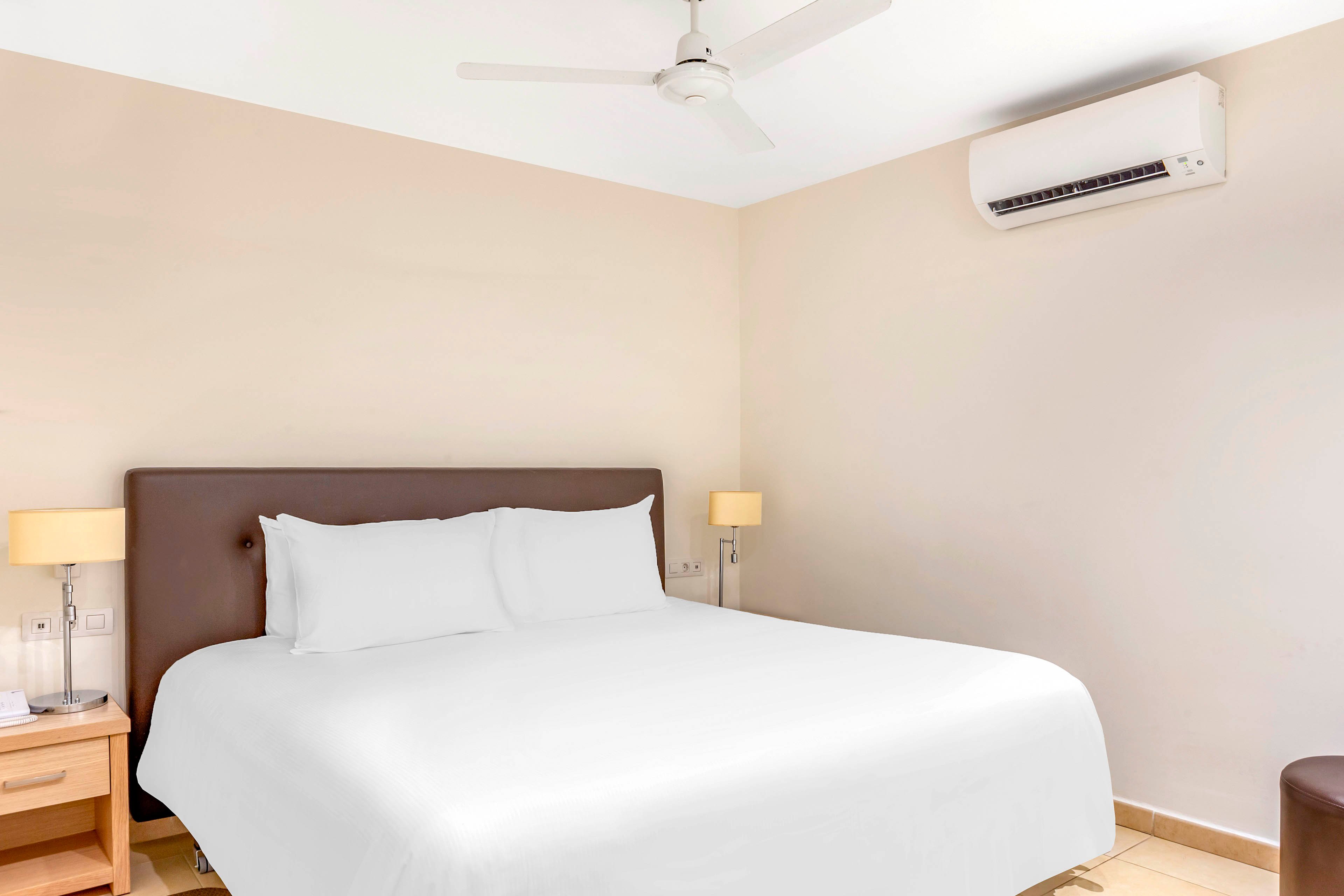 A neatly made double bed with crisp white bedding stands against a light-colored wall in a minimalist bedroom. Two bedside tables with matching lamps flank the bed, adding symmetry to the space. A ceiling fan and wall-mounted air conditioner suggest a comfortable, climate-controlled environment. The neutral color palette and uncluttered decor create a calm, modern atmosphere.