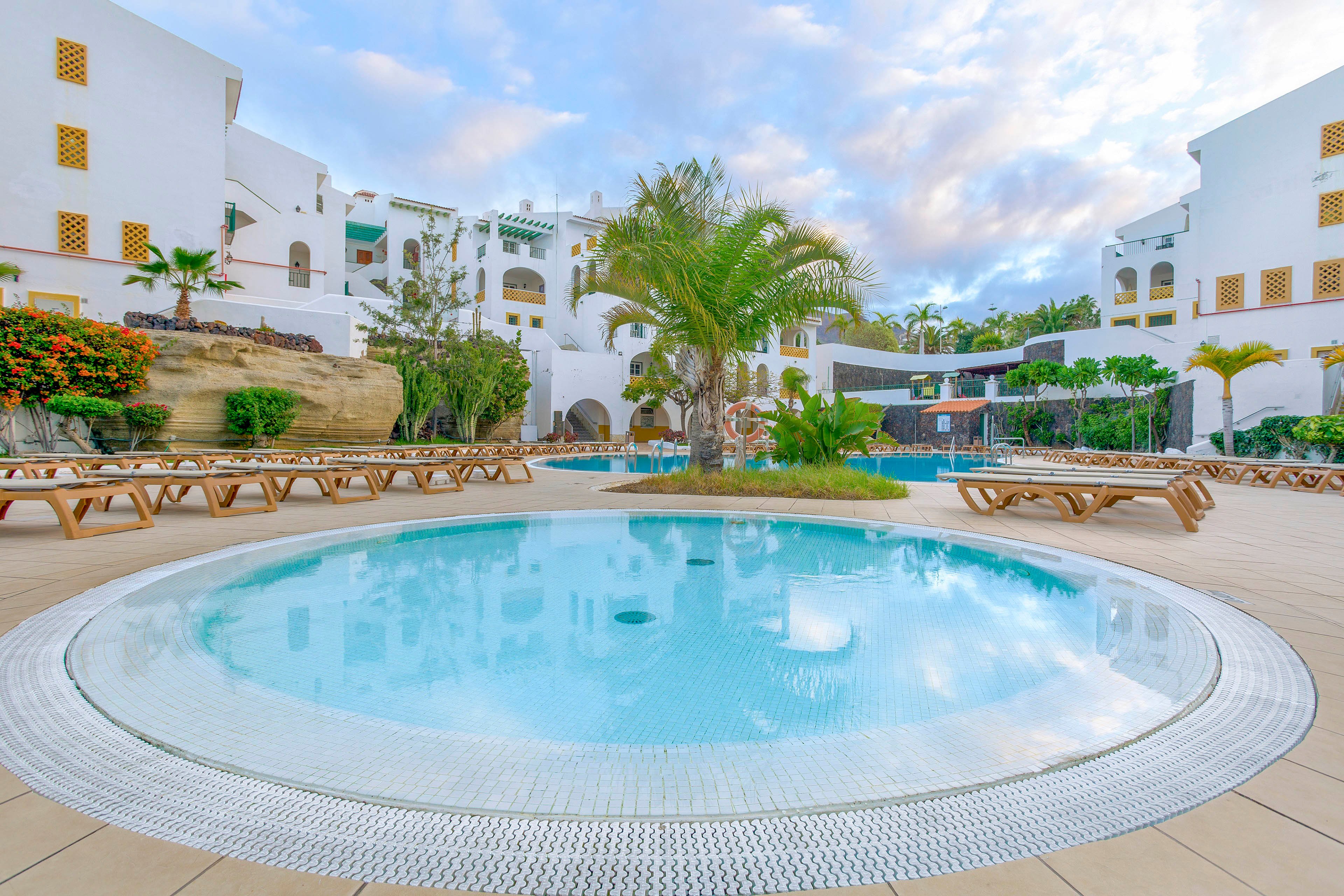 A circular shallow pool sits at the center of a resort courtyard. White Mediterranean-style buildings with arches and balconies surround the pool area. Wooden sun loungers line the tiled deck, and a tall palm tree adds a tropical accent. The sky is partly cloudy, creating a soft, relaxed atmosphere ideal for vacation leisure.