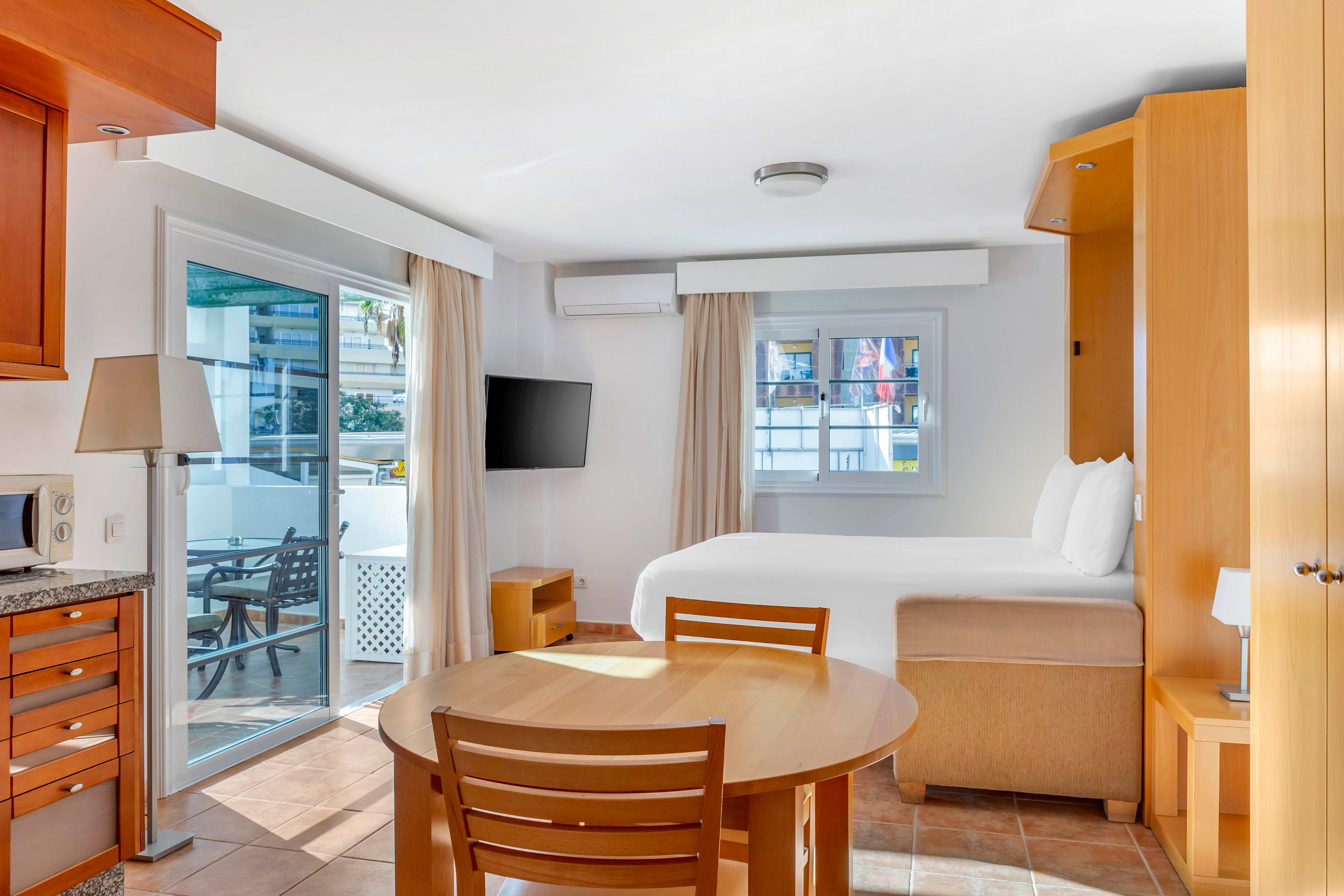 A compact studio-style hotel room features a neatly made double bed beside a large window. A round wooden dining table with chairs sits in the foreground, facing a wall-mounted flat-screen TV. Sliding glass doors open to a sunny balcony with outdoor seating and views of neighboring buildings. Warm wood cabinetry, tiled floors, and neutral curtains create a clean, inviting atmosphere.