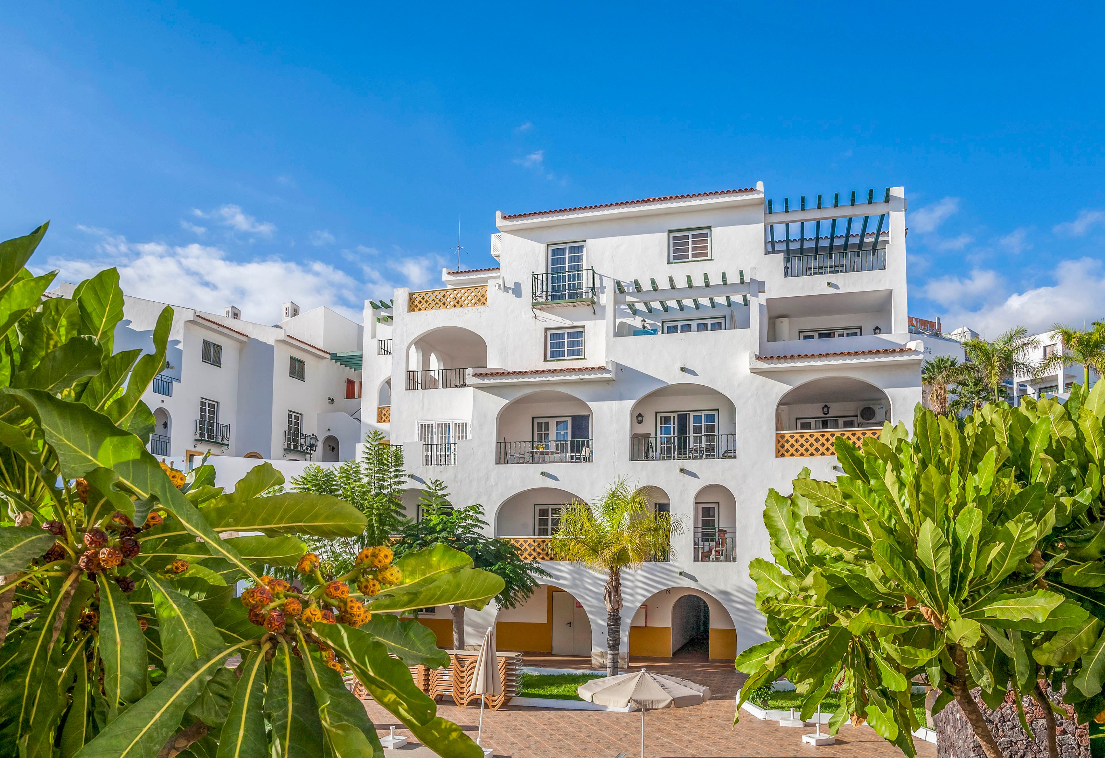A white multi-story apartment building with arched balconies stands under a clear blue sky. The architecture features Mediterranean influences with terracotta roof tiles and decorative railings. Lush green tropical plants and palm trees frame the foreground, adding vibrant color and texture. Sunlight enhances the bright, clean facade, creating a warm and inviting holiday atmosphere.