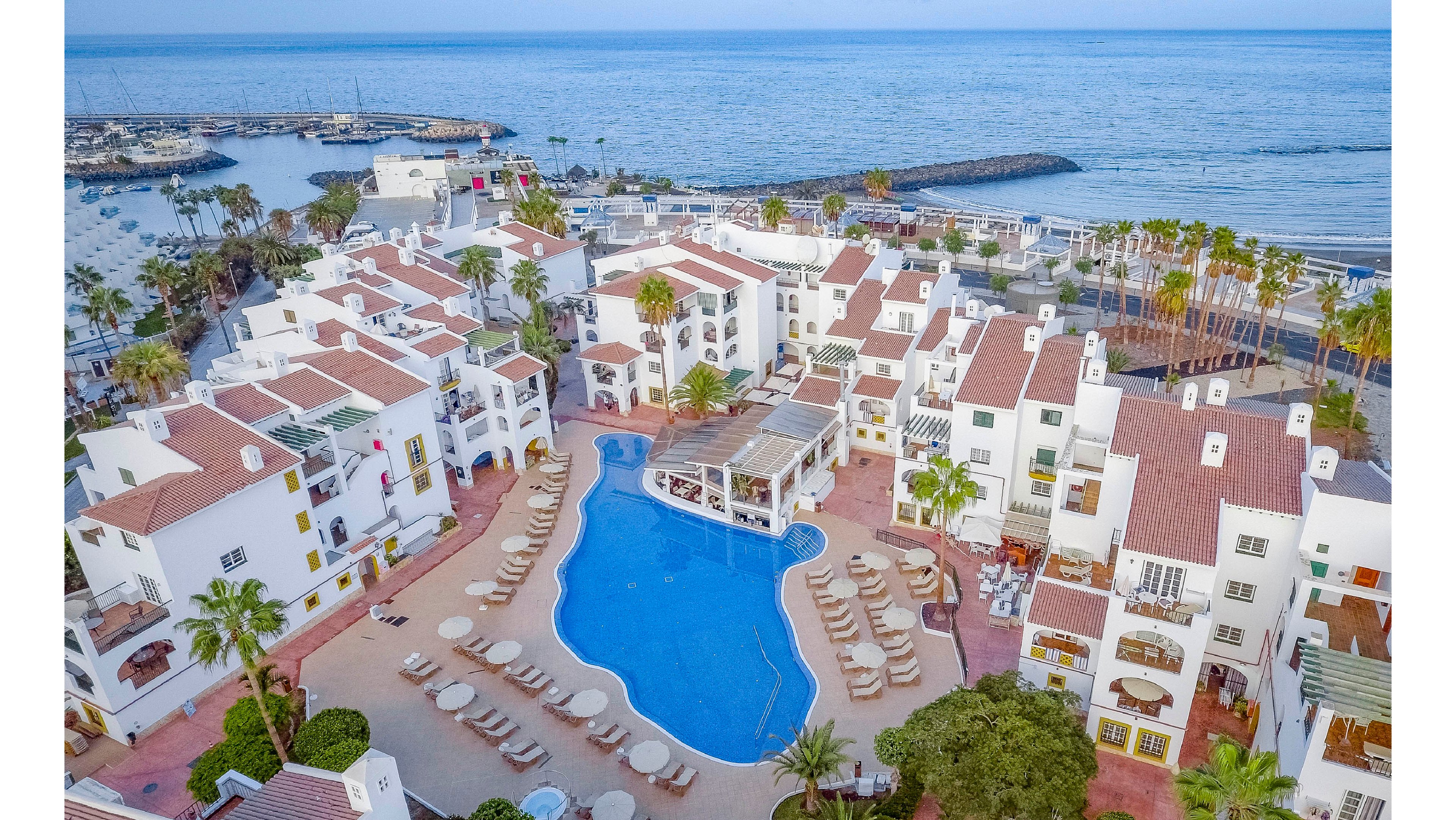 A large seaside resort complex features a central blue swimming pool surrounded by sun loungers and umbrellas. White multi-story buildings with red-tiled roofs encircle the pool, creating a courtyard atmosphere. Palm trees and landscaped walkways add a tropical feel to the scene. In the background, the calm ocean and a small harbor with breakwaters extend to the horizon under a clear sky.