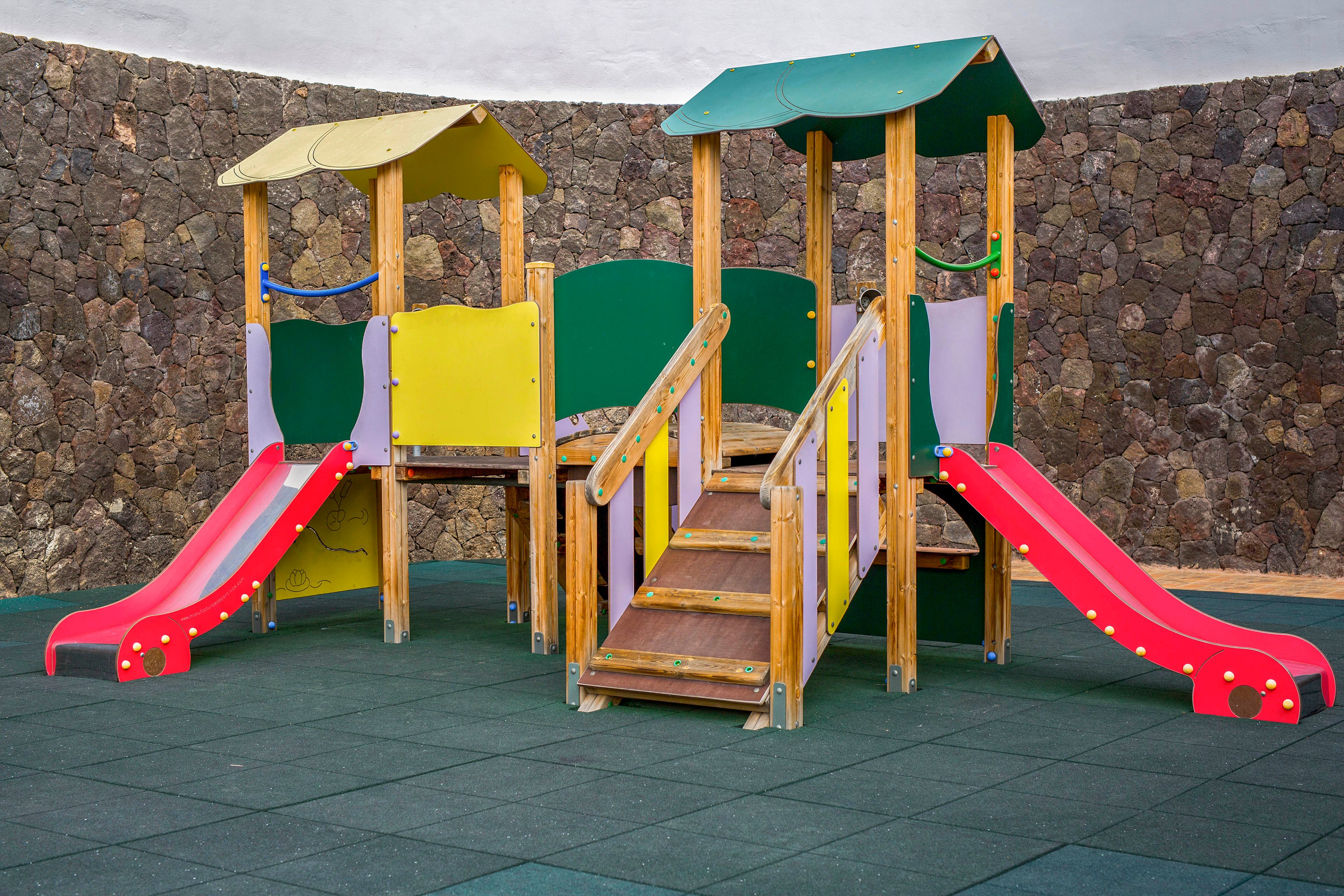 A brightly colored wooden playground structure stands on a rubber safety surface. The playset features multiple platforms, stairs, and several red slides. Curved roofs and side panels in yellow, green, and purple add a cheerful appearance. A high stone wall encloses the quiet outdoor play area, with no children present.