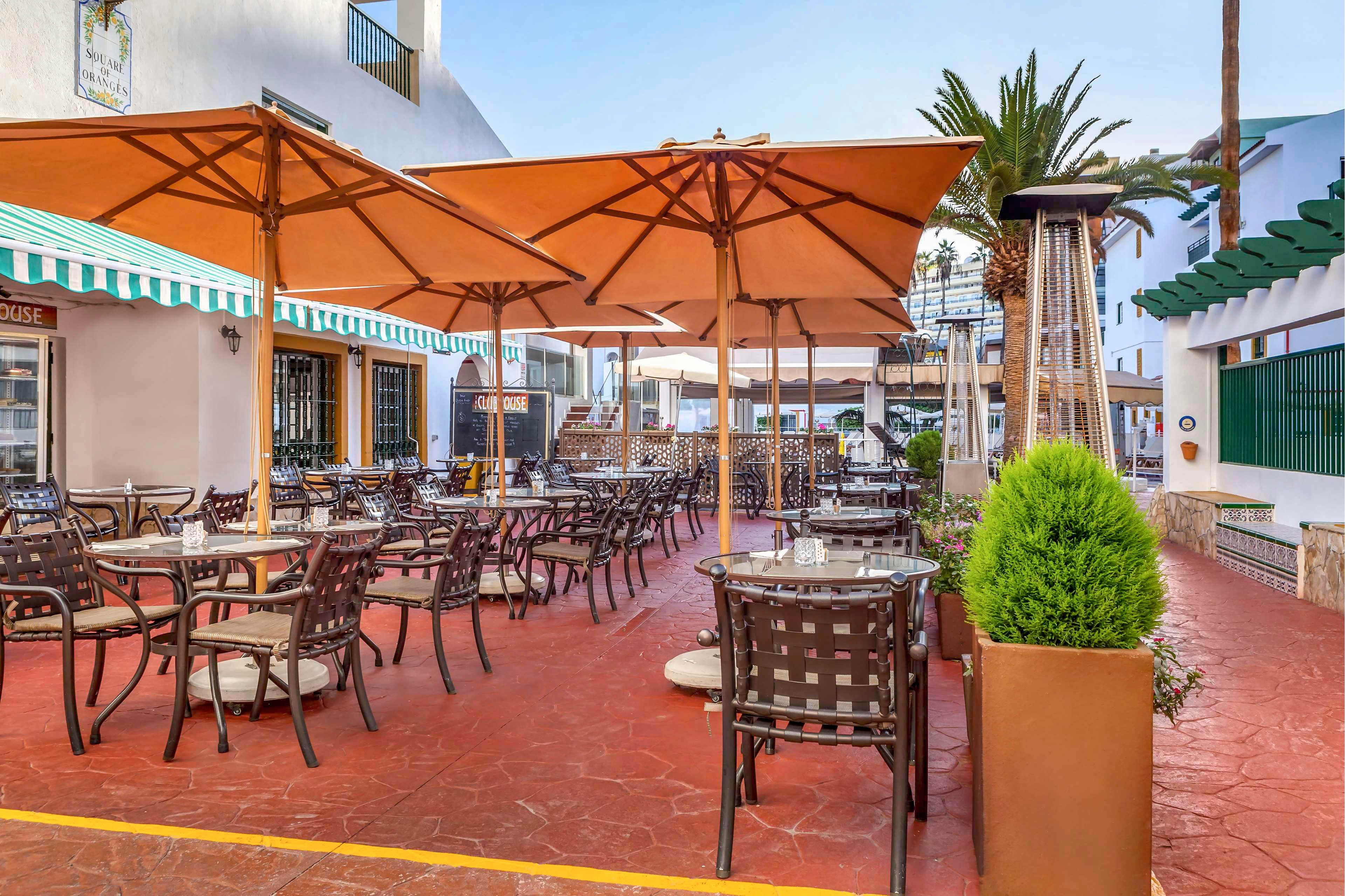 An open-air cafe terrace features numerous tables and chairs neatly arranged under large beige umbrellas. The setting appears to be in a warm climate courtyard, surrounded by low white buildings with striped awnings and green accents. A tall outdoor heater and potted plants add comfort and greenery to the space. The scene is bright and inviting, suggesting a relaxed atmosphere for dining or socializing.