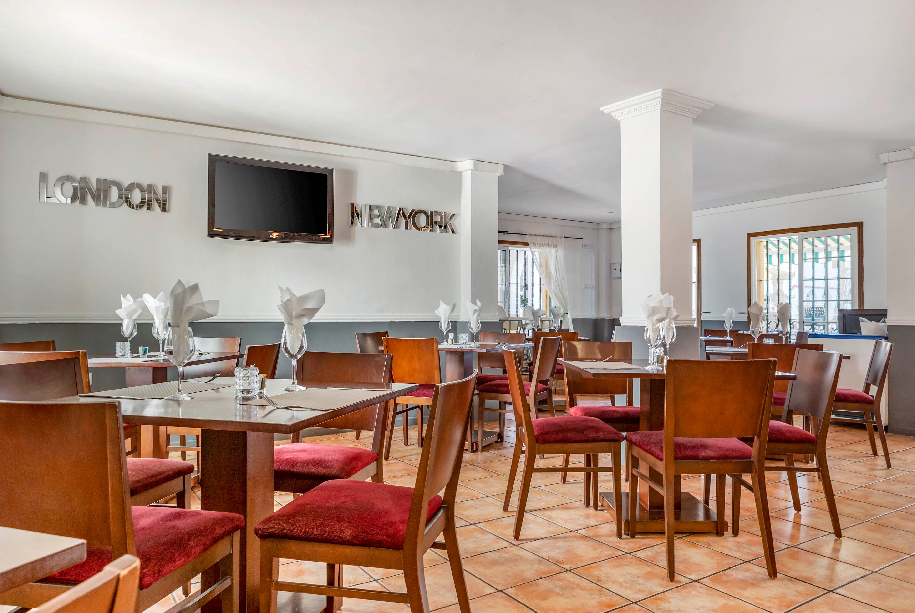Contemporary restaurant dining area with wooden tables and red cushioned chairs is shown in a bright, open space. Tables are neatly set with folded white napkins, glassware, and cutlery, suggesting readiness for guests. A flat-screen television is mounted on the wall between metallic wall text reading LONDON and NEW YORK. Large windows and neutral walls create an airy, welcoming atmosphere with tiled flooring and clean architectural lines.