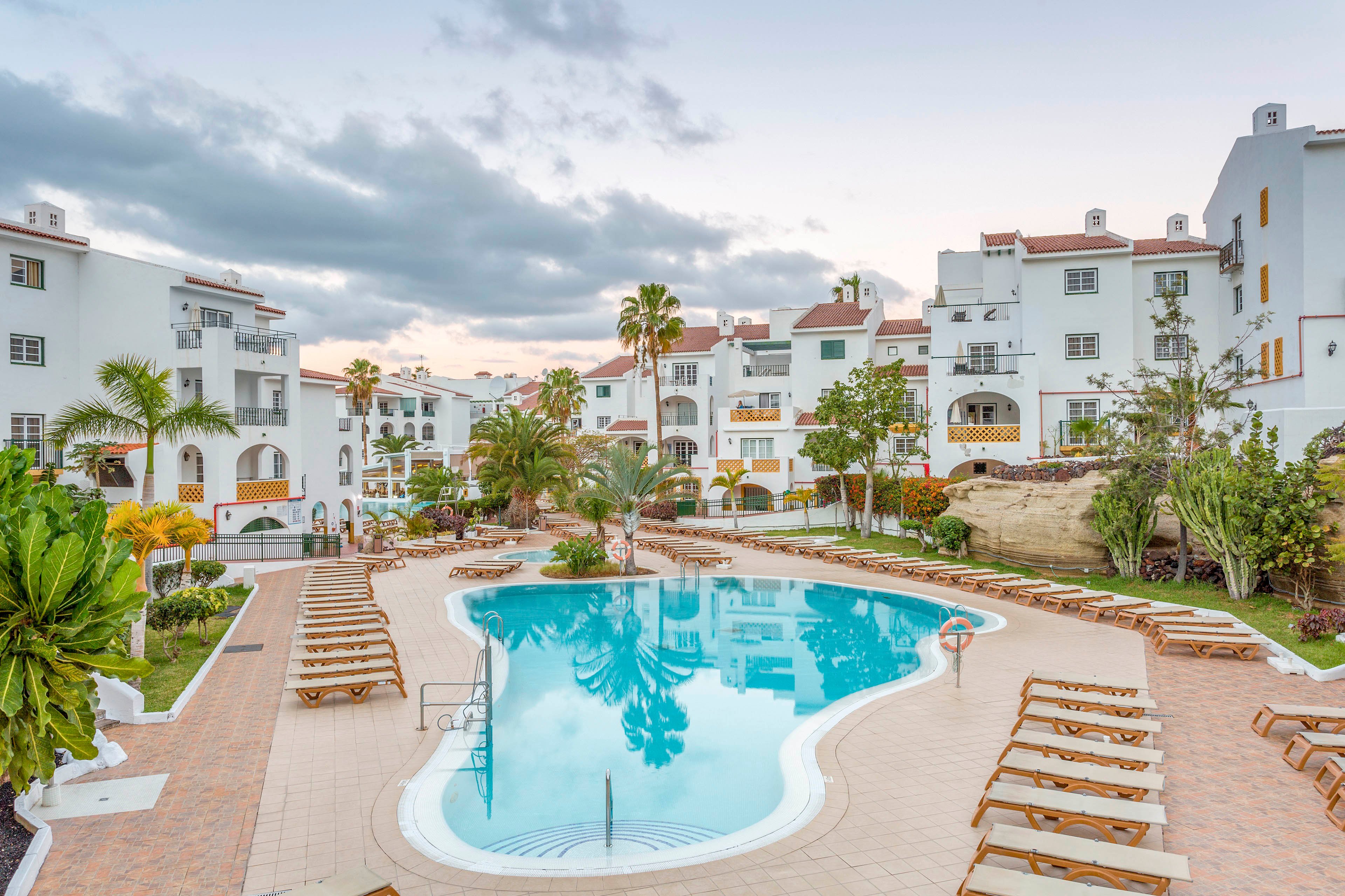 A tranquil resort courtyard features a large, curved outdoor swimming pool surrounded by sun loungers. White multi-story apartment-style buildings with red roofs frame the pool area, creating a Mediterranean or coastal village feel. Palm trees, lush greenery, and landscaped pathways add to the vacation atmosphere. The sky is overcast with soft clouds, giving the scene a calm, relaxed mood.