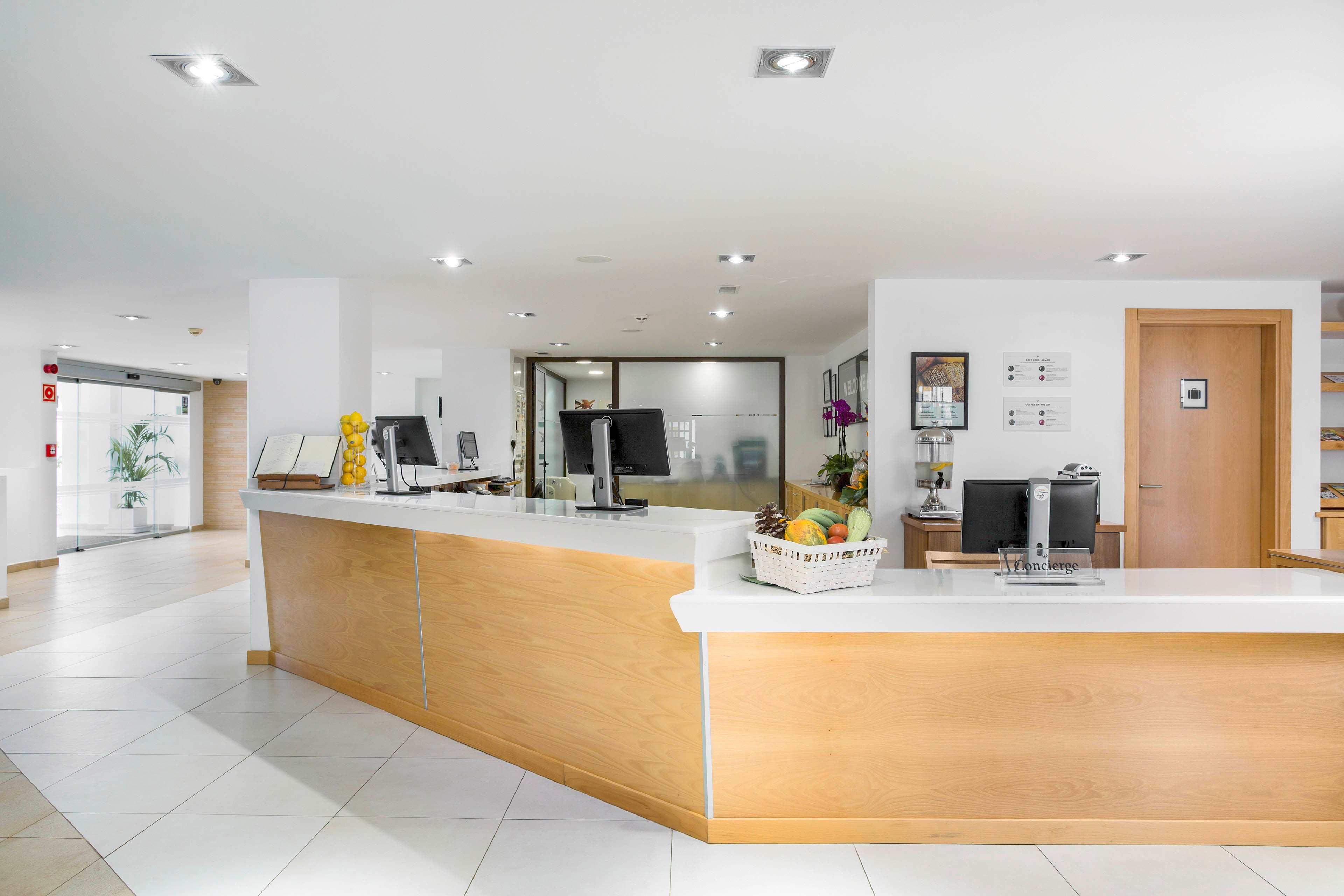 A spacious reception area features a large wooden front desk with a white countertop. Computer monitors, office supplies, and a basket of fruit sit neatly on the counter. The lobby is bright and minimalist, with white walls, tiled floors, and recessed ceiling lights. Doors, glass partitions, and wall-mounted frames are visible in the background, enhancing the clean, professional atmosphere.