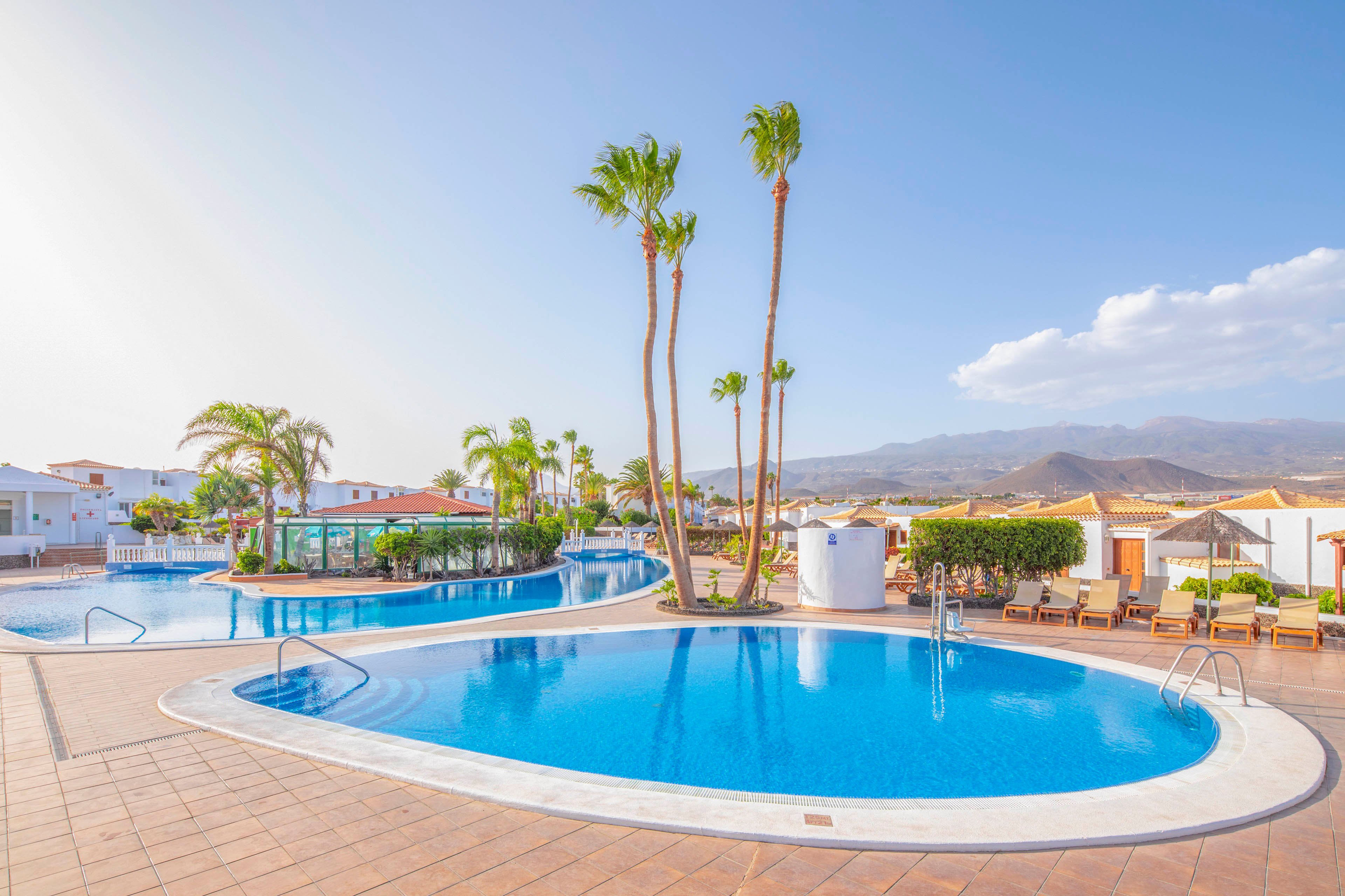 A bright resort swimming pool curves through a spacious outdoor terrace under a clear blue sky. Tall palm trees rise from the pool area, adding a tropical feel to the scene. Sun loungers and umbrellas line the deck, suggesting a relaxing vacation atmosphere. Low white buildings and distant mountains frame the background, enhancing the sense of openness and leisure.