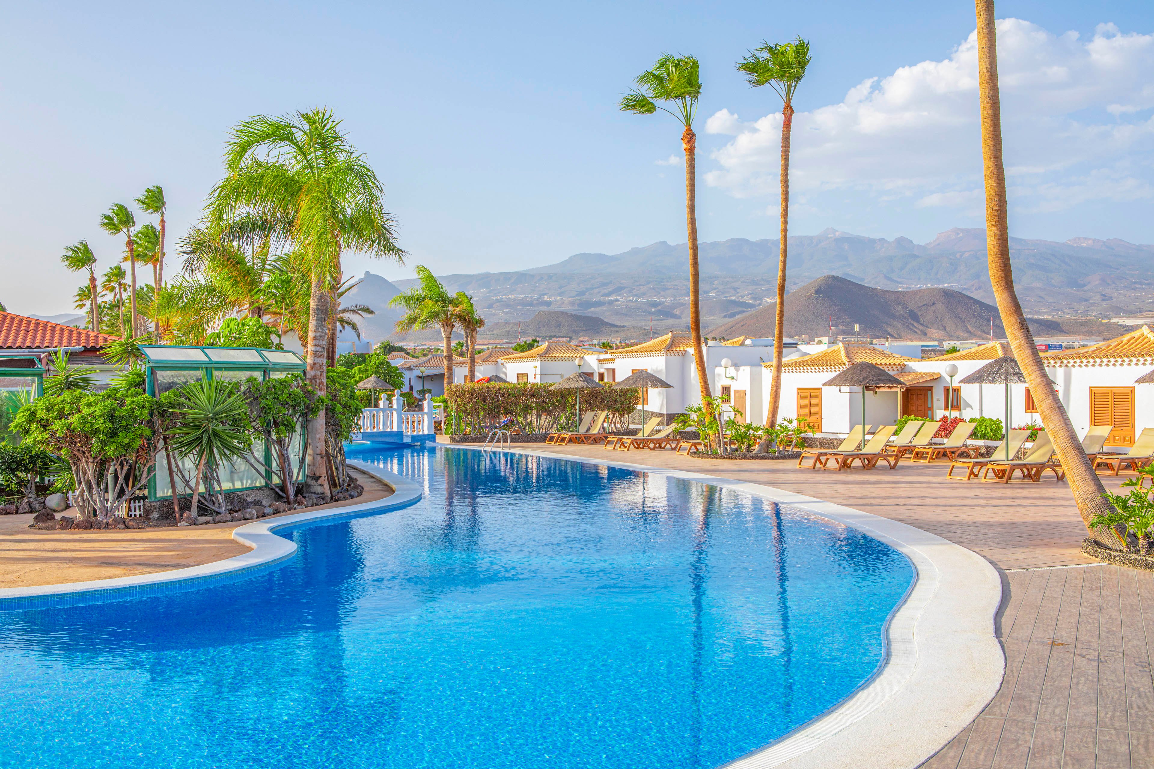 Outdoor resort swimming pool curves through a landscaped terrace lined with palm trees and sun loungers. White bungalows with red-tiled roofs border the pool area, creating a relaxed vacation setting. Clear blue water reflects the bright sky and distant mountains in the background. The scene suggests a warm, tranquil holiday destination ideal for sunbathing and leisure.