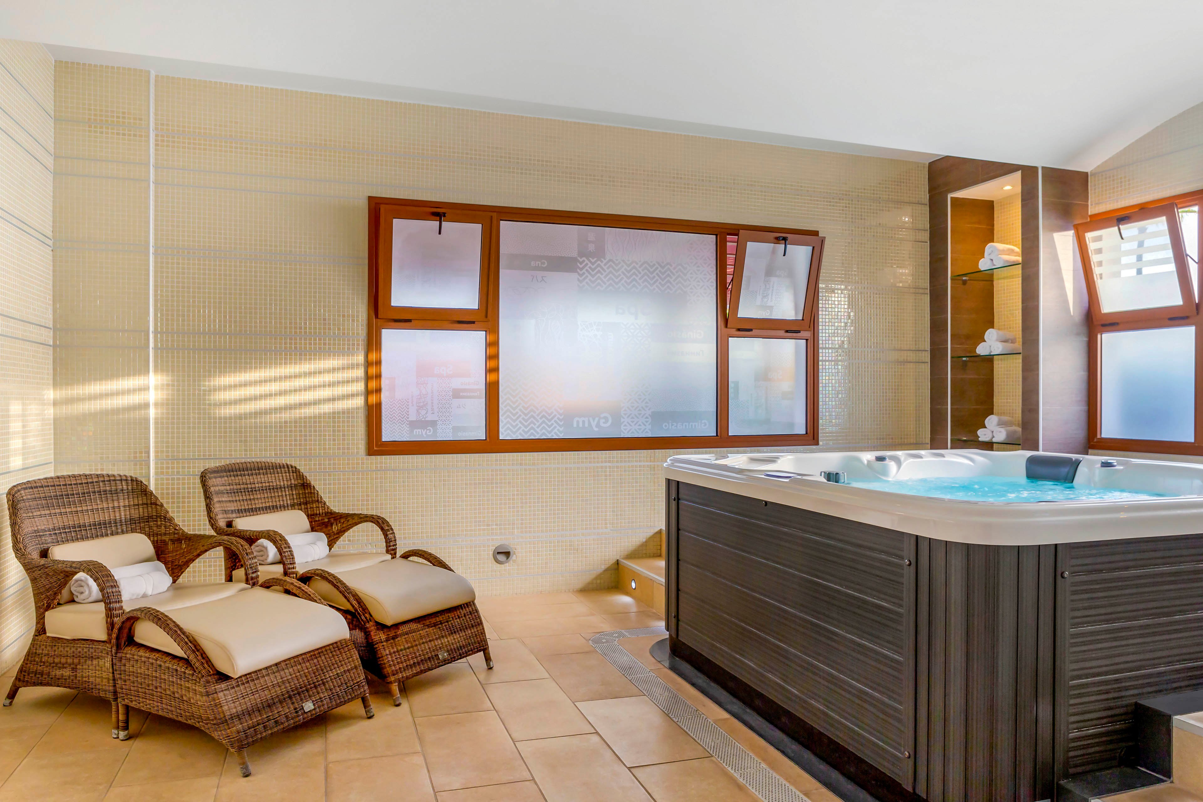 A contemporary spa room features a large built-in hot tub as the central focus. Two wicker lounge chairs with cushions and rolled towels are arranged beside the tub for relaxation. Frosted windows with wooden frames allow natural light to filter into the beige-tiled space. Open shelving in the corner holds neatly folded towels and spa accessories, creating a calm and inviting atmosphere.