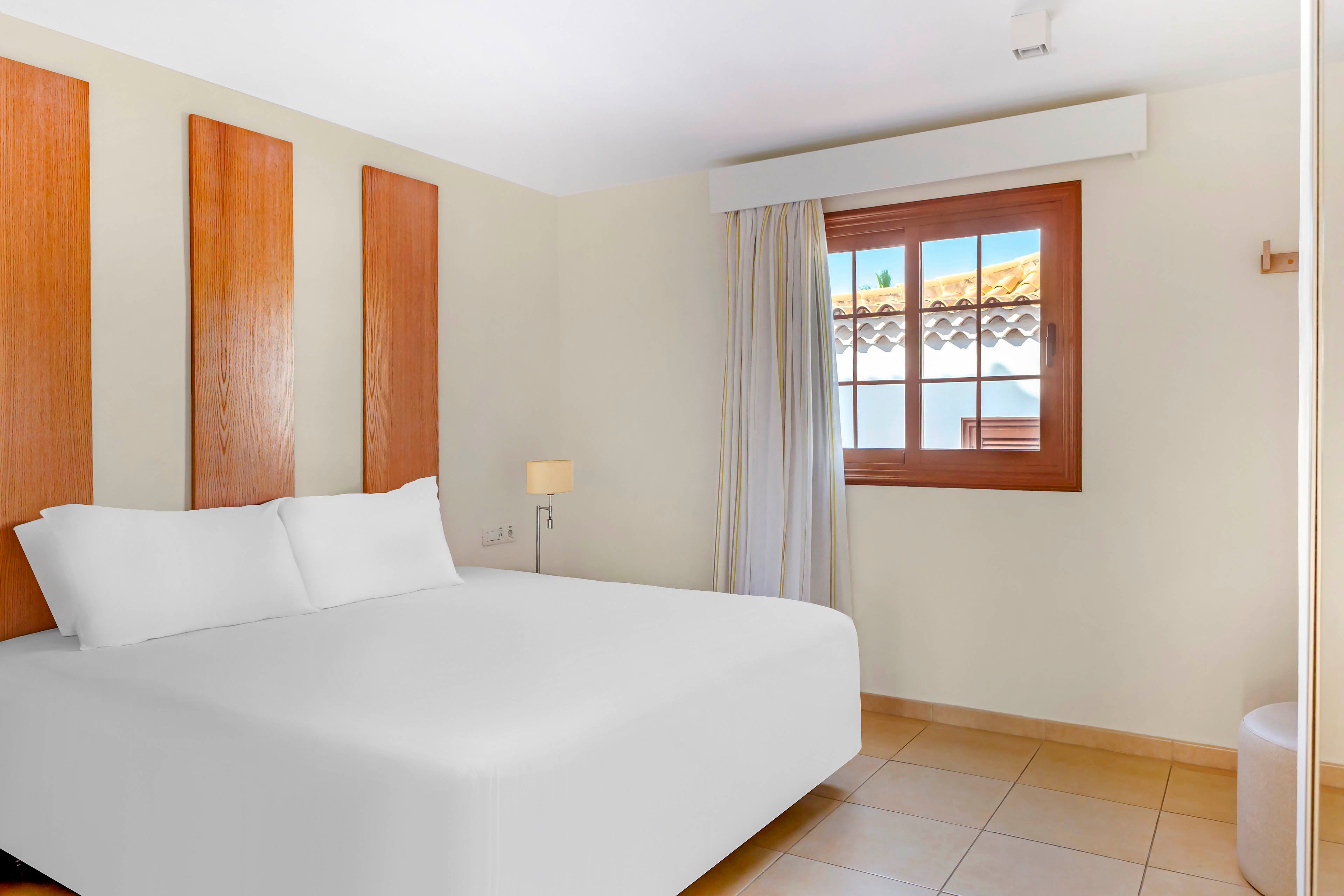 A clean, minimalist bedroom features a large neatly made bed with white linens. The room includes light-colored tiled flooring, cream walls, and three vertical wooden panels as decorative elements above the bed. A small bedside lamp and curtain-covered window add warmth and natural light, with a tiled rooftop visible outside. The overall atmosphere is calm, modern, and inviting, suitable for a hotel or vacation rental.