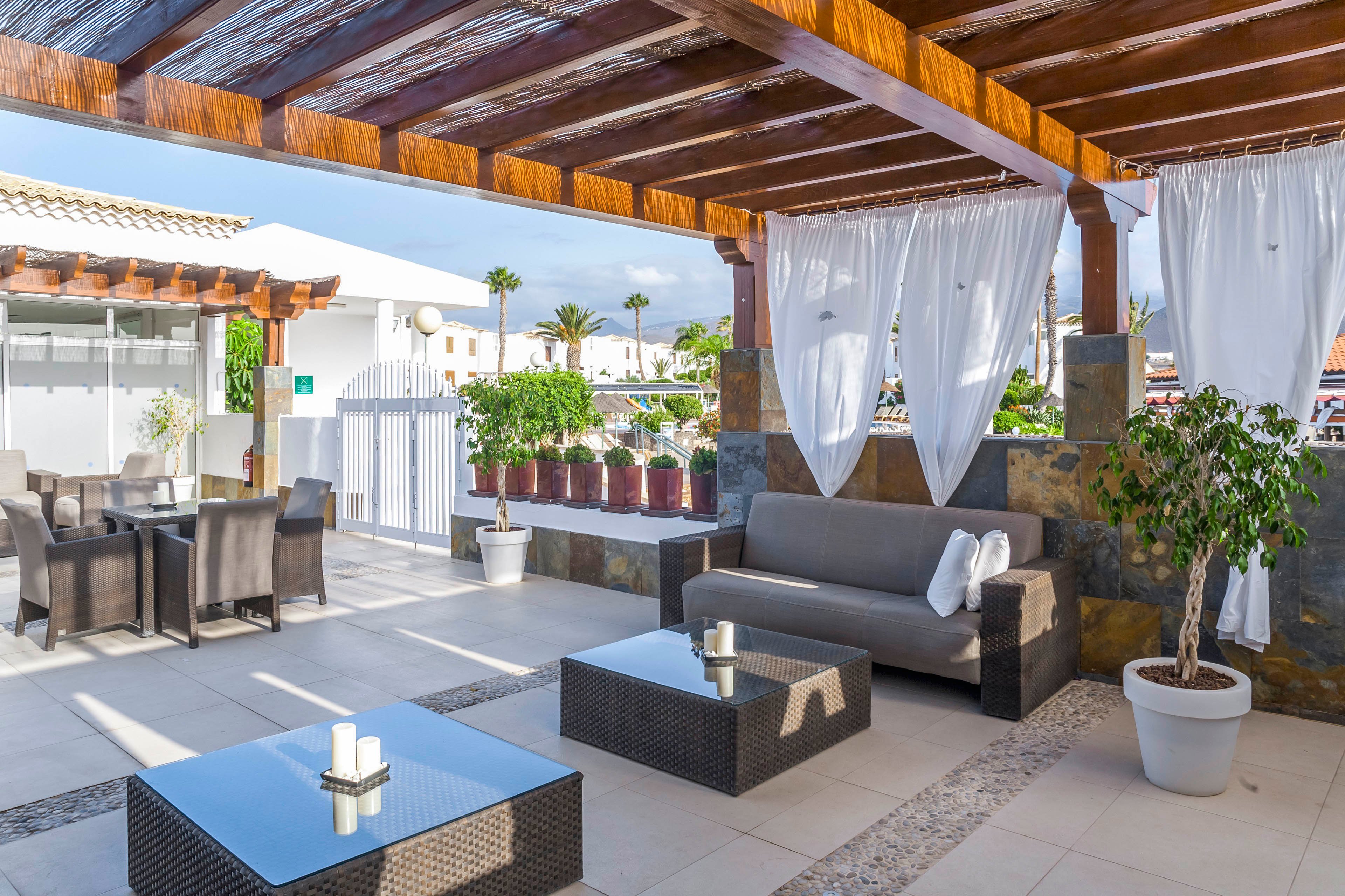 Contemporary outdoor lounge area features a wooden pergola casting shadows over sleek wicker furniture. A gray sofa with white towels and low coffee tables sit on a tiled terrace. White curtains, potted plants, and decorative stones add a resort-like ambiance. In the background, additional seating and white buildings suggest a sunny vacation setting.