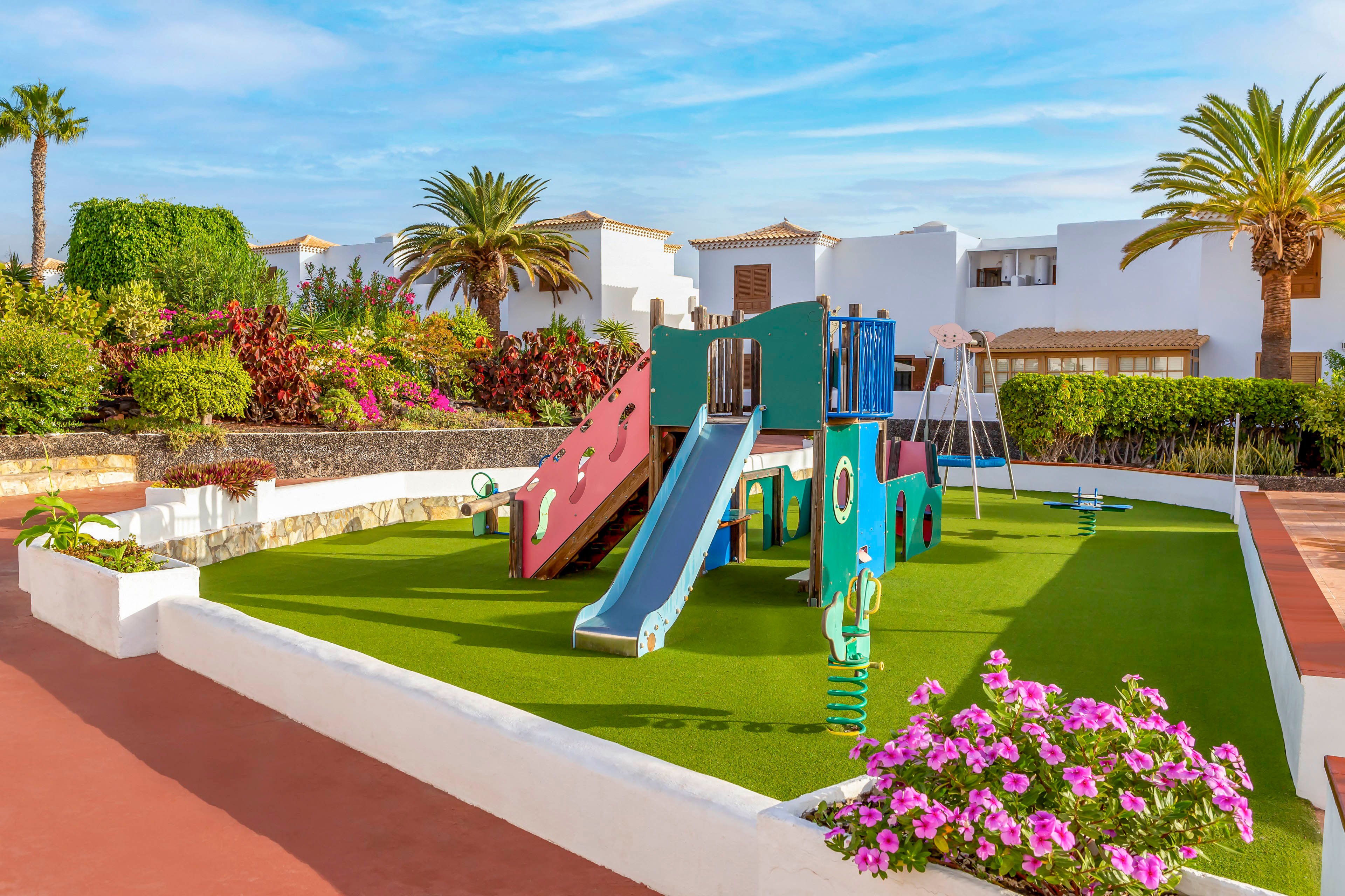 A small colorful playground with slides and climbing structures sits on a bright green artificial grass surface. It is located in an outdoor courtyard surrounded by white low-rise buildings and lush tropical plants. Pink flowers border the play area, adding vibrant contrast to the scene. The clear blue sky and palm trees create a warm, inviting holiday atmosphere.