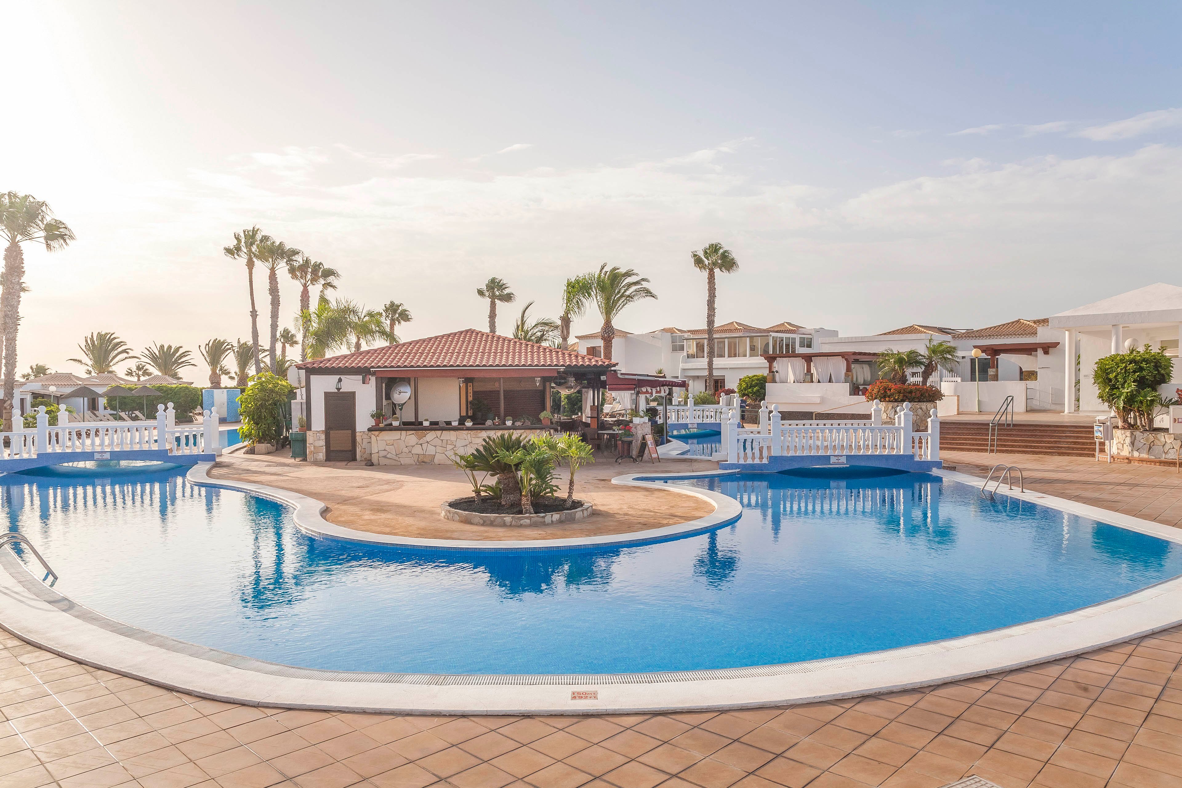 A large, curved swimming pool dominates a sunny resort scene. In the center, a small poolside bar with a red-tiled roof sits on an island surrounded by clear blue water. White railings, palm trees, and low vacation villas frame the pool area. The warm light and calm water create a relaxed, holiday atmosphere.