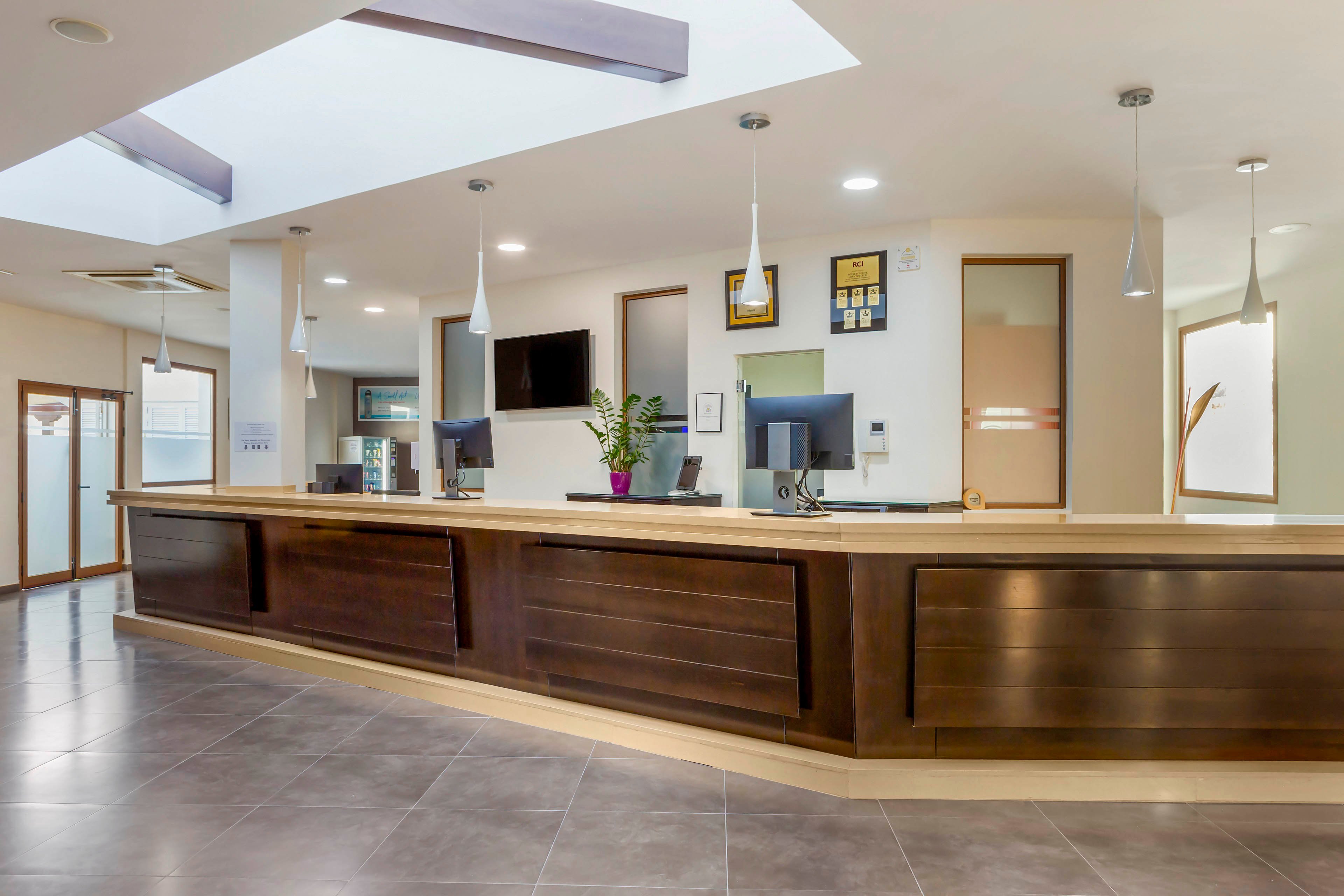 A long wooden reception desk dominates a spacious, modern lobby area. Computer monitors, a telephone, and a potted plant sit neatly on the countertop. Pendant lights hang from the ceiling, illuminating the clean, minimalist interior. Framed certificates and artwork decorate the walls behind the desk, while large windows and glass doors bring in natural light.