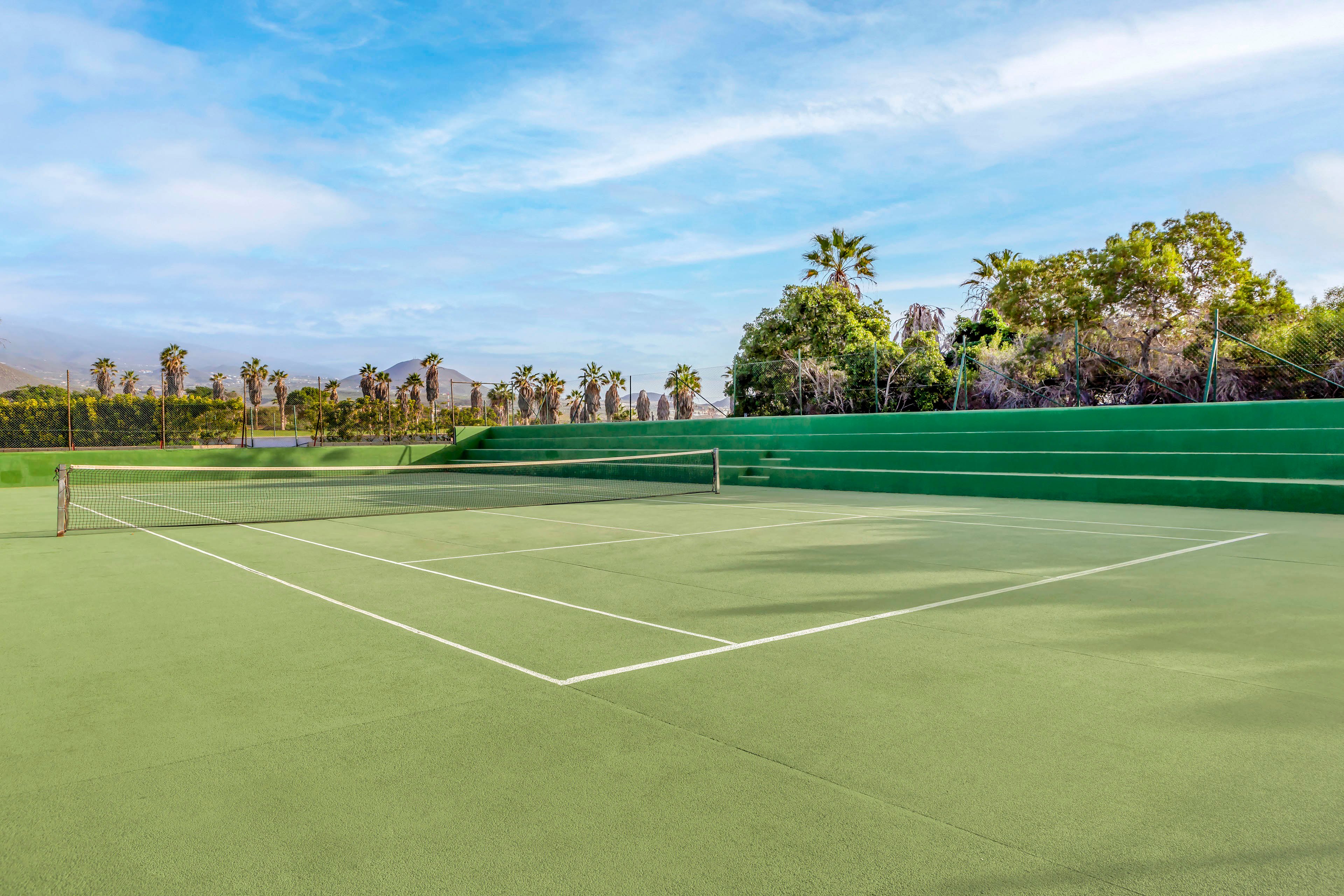 An empty outdoor tennis court is shown under a bright blue sky with light clouds. The green hard court surface and white boundary lines are clearly visible. Surrounding the court are green walls, trees, and palm trees, with distant hills or mountains in the background. The scene feels calm and quiet, suggesting early morning or off-peak playtime.