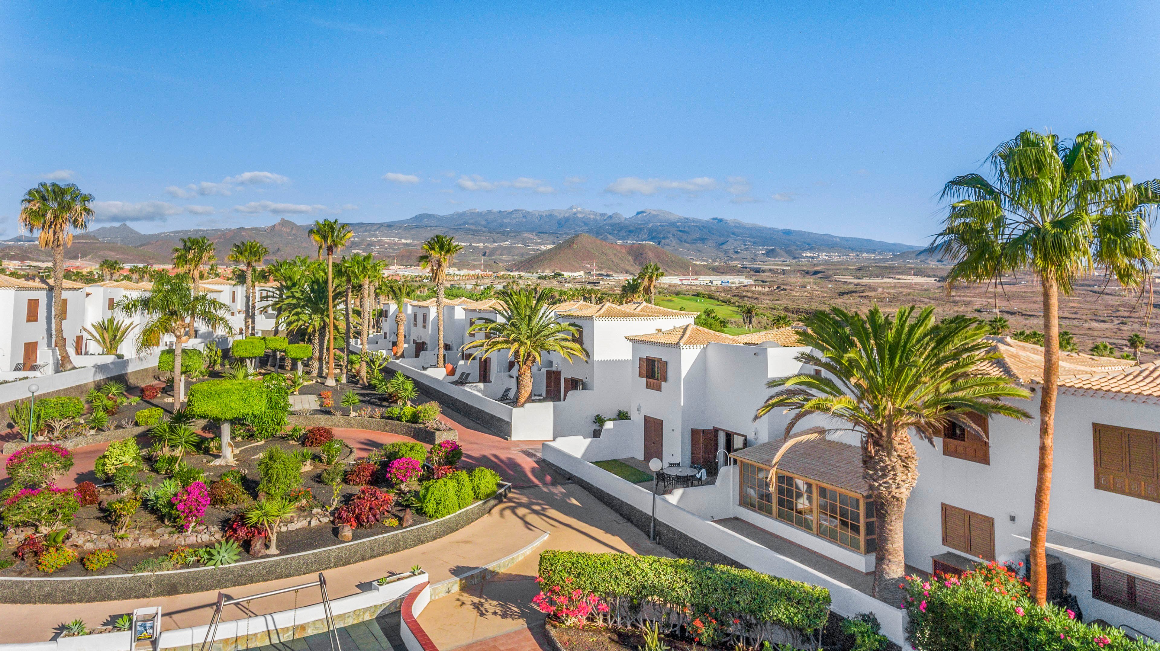 A bright resort complex features white, flat-roofed buildings surrounding a landscaped courtyard. Palm trees, trimmed hedges, and colorful flowering plants create a lush garden in the center. In the background, arid hills and distant mountains stretch under a clear blue sky. The scene suggests a warm, dry climate and a peaceful vacation setting.