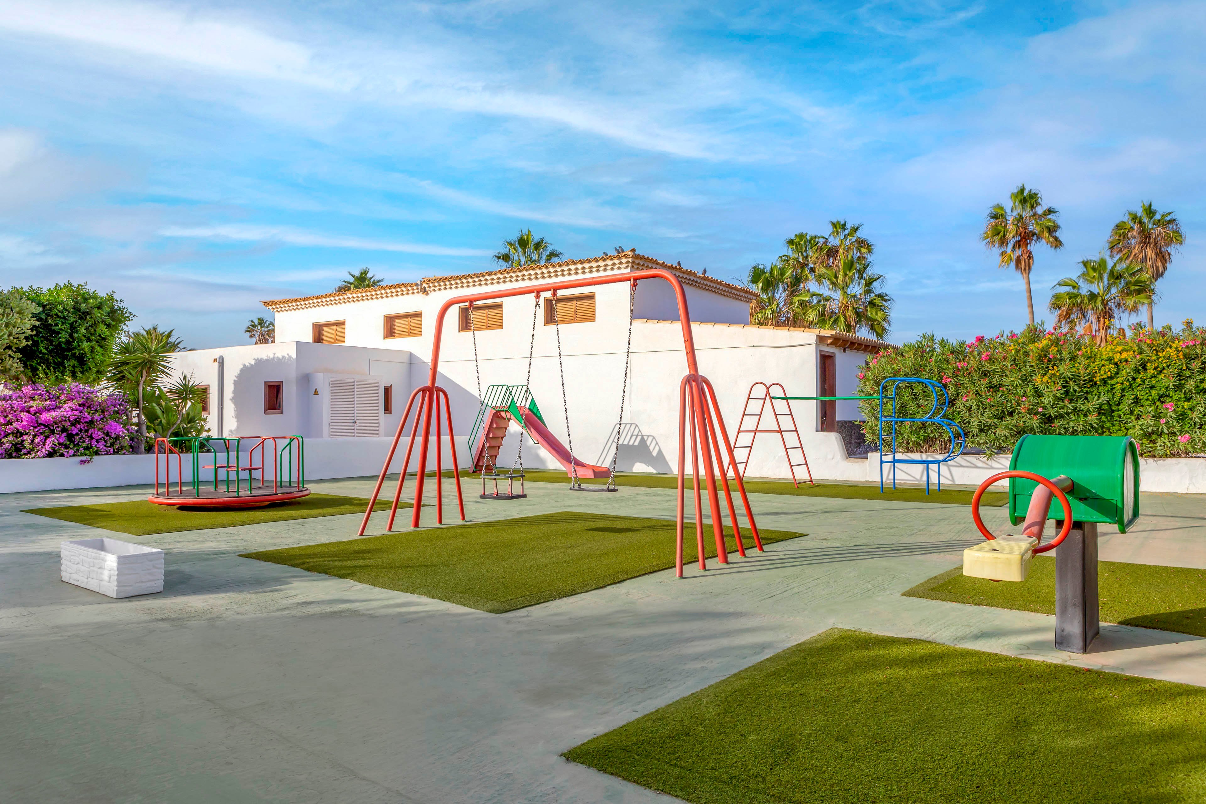 An empty outdoor playground with colorful metal swings and climbing frames sits in front of a white villa. The play area features artificial grass islands set into a smooth concrete surface. Bright flowers and green hedges border the space, with tall palm trees rising behind the house. The sky is clear and blue, suggesting a sunny, warm day in a residential or vacation setting.