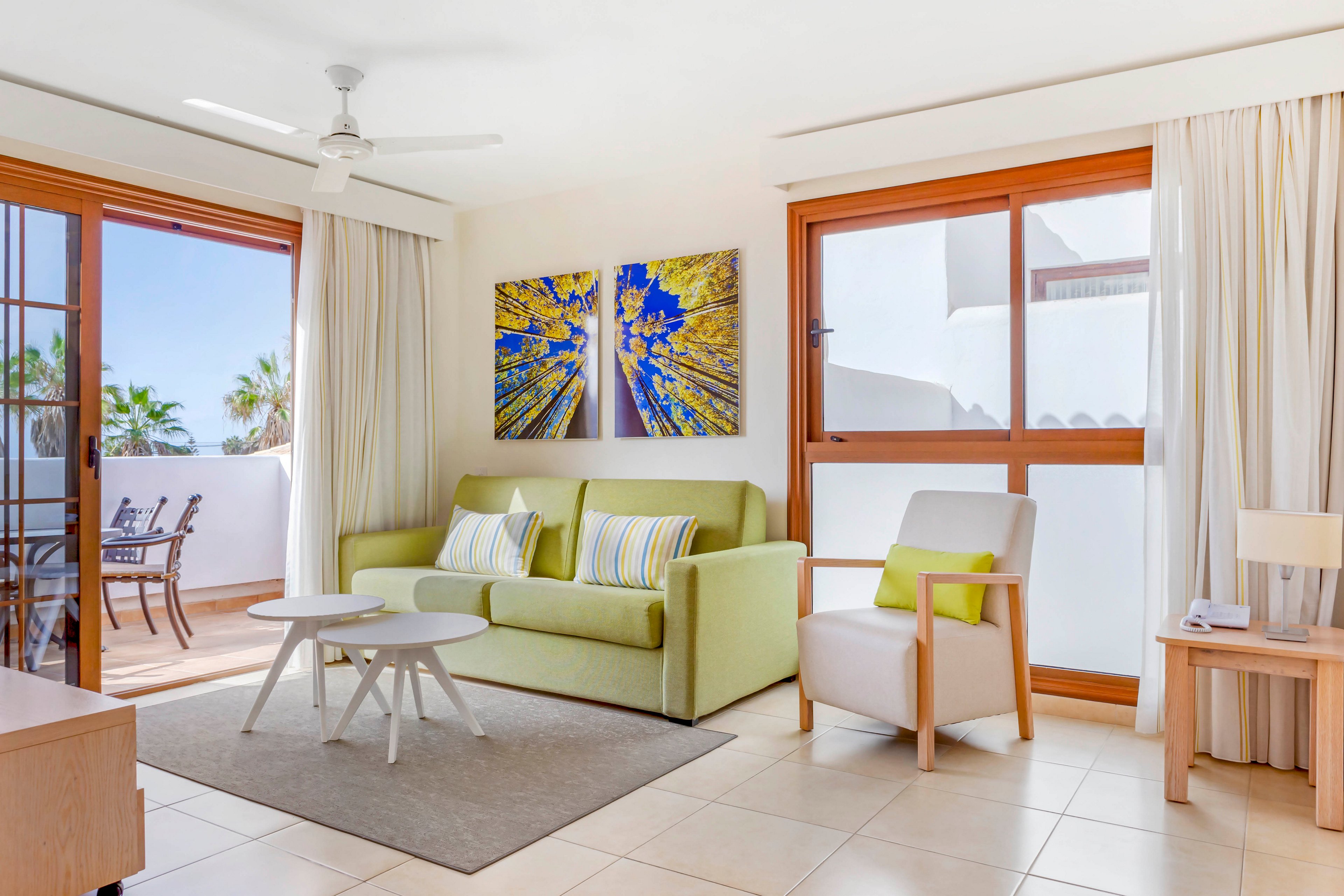 A contemporary living room features a light green sofa and a white armchair with colorful cushions. Large windows and glass doors let in abundant natural light and open to a balcony with outdoor seating. Minimalist furniture, including a round coffee table and side tables, sits on a neutral rug over tiled flooring. White walls, curtains, and ceiling fan contribute to the airy, vacation-like atmosphere, complemented by vibrant abstract artwork on the wall.