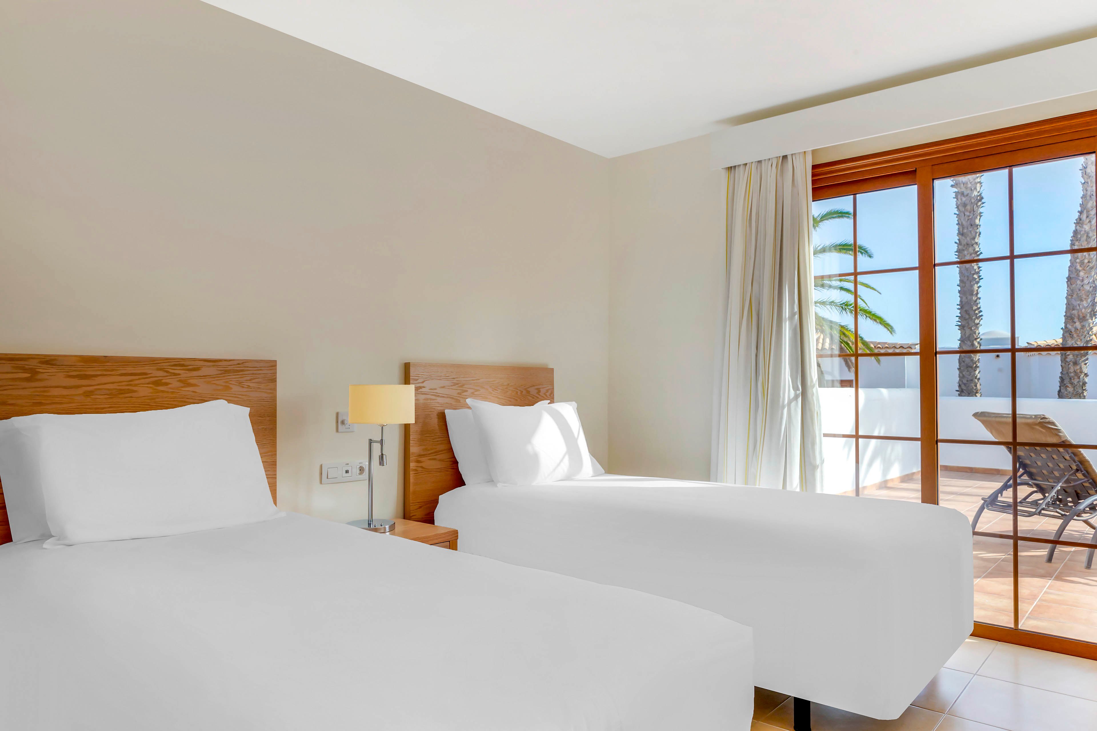 A modern hotel room features two neatly made twin beds with white linens and wooden headboards. A bedside table with a lamp sits between the beds against a neutral-colored wall. Large sliding glass doors open to a sunny terrace with outdoor chairs and palm trees visible outside. Natural light fills the room, creating a clean and inviting atmosphere.