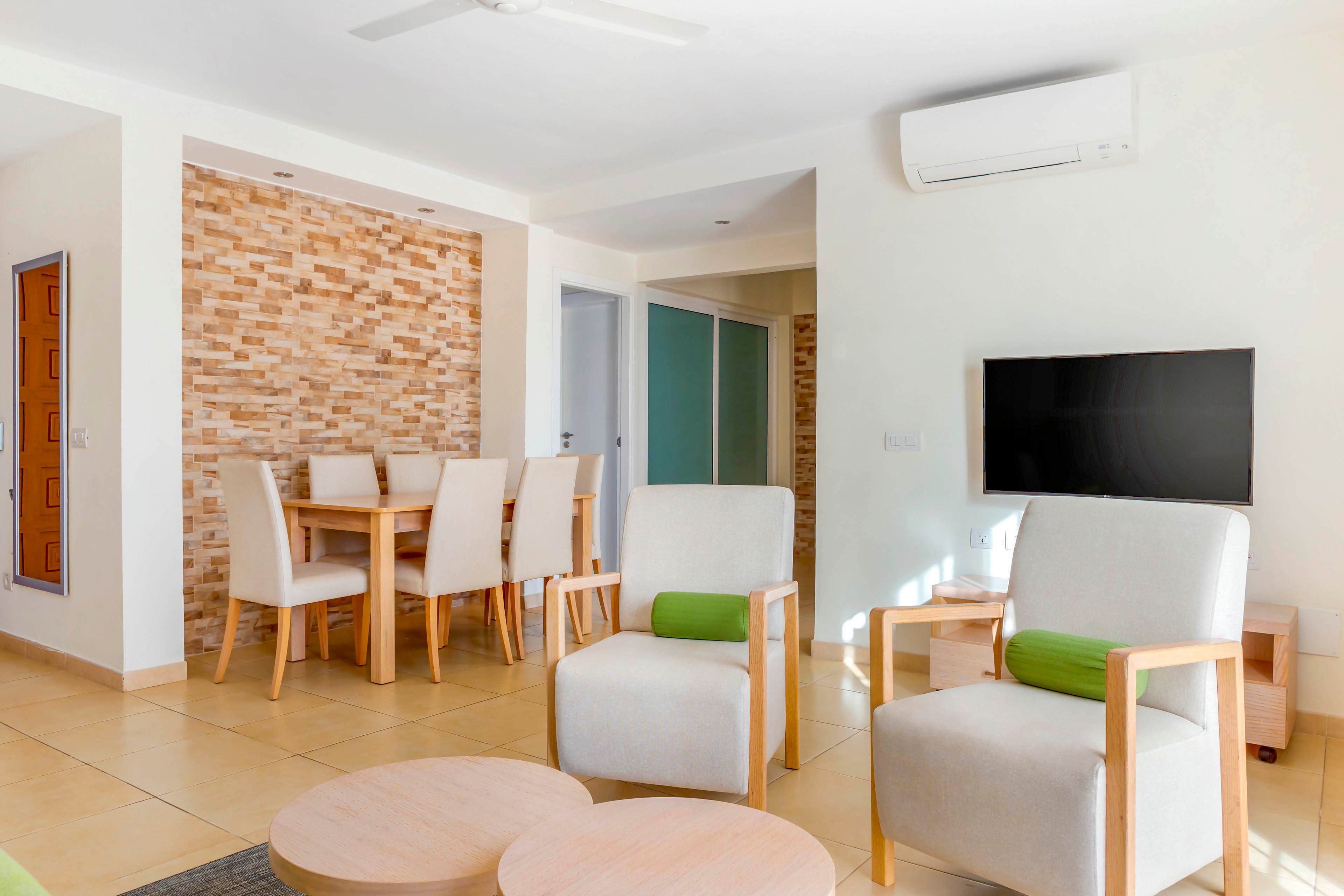 Contemporary living room interior features light wooden armchairs with green cushions and round coffee tables. A flat-screen television is mounted on the wall beneath a white air conditioning unit. In the background, a dining area with a wooden table and upholstered chairs sits against a textured brick accent wall. The space is clean, minimal, and filled with natural light, emphasizing a neutral color palette with subtle green accents.