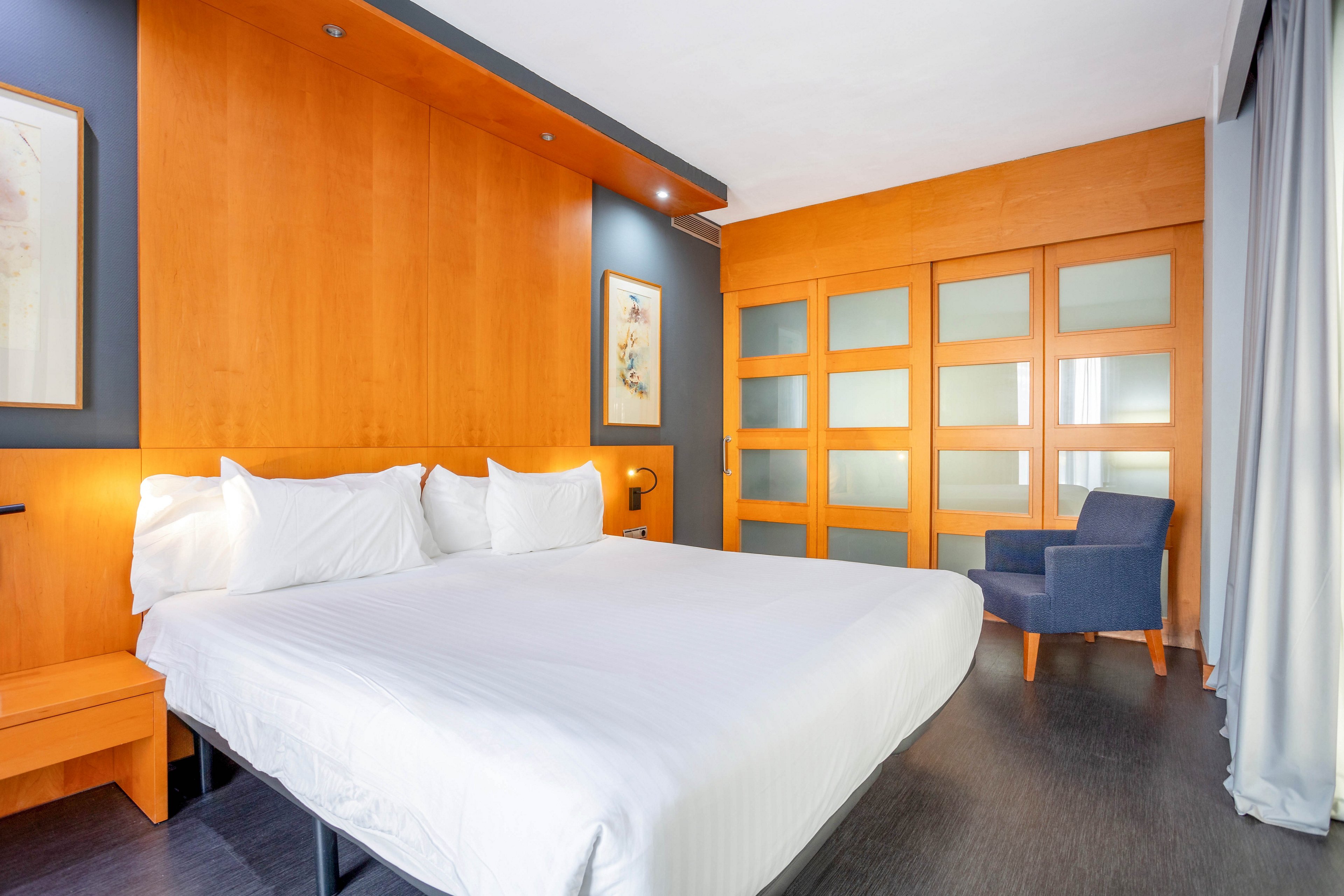 A spacious hotel room features a large king-sized bed with crisp white linens and pillows. The setting includes warm wood paneling, frosted glass sliding doors, and a comfortable blue armchair by the window. The room is well-lit, with minimalist decor and a neutral palette, creating a clean and inviting atmosphere. No visible text or numeric values are present in the image.