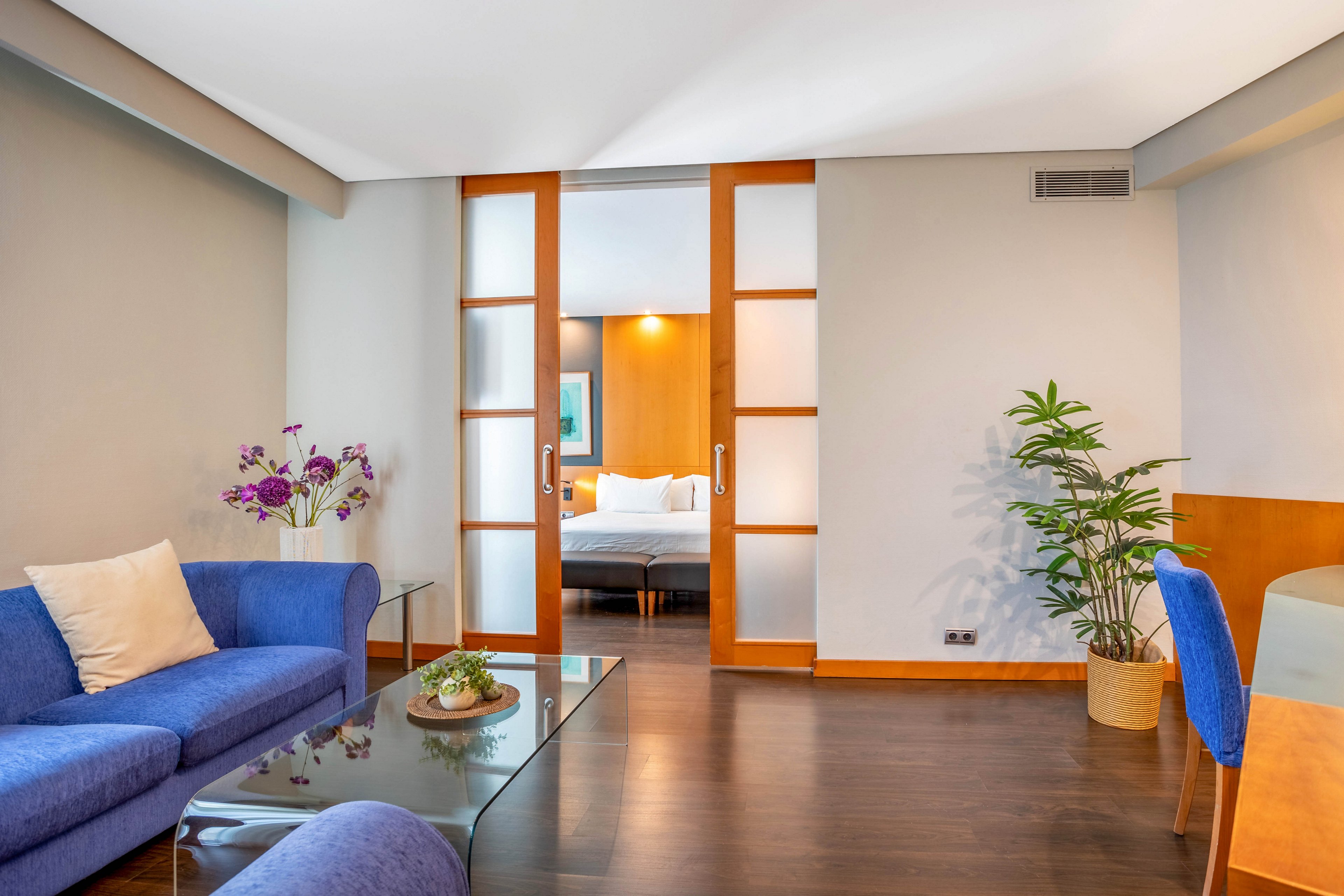 A contemporary hotel suite features a blue sofa with a white pillow, a glass coffee table, and decorative flowers. Sliding wooden doors open to reveal a bedroom with a neatly made bed and soft lighting. The room is accented with indoor plants and minimalist decor, creating a calm and inviting atmosphere.
