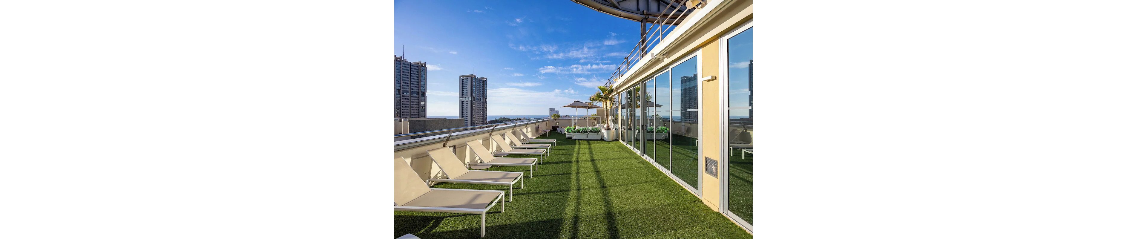 A spacious rooftop terrace features several beige lounge chairs arranged on artificial green grass. The setting is urban, with tall buildings visible in the background and a clear blue sky overhead. Glass windows reflect the outdoor scene, and potted plants add a touch of greenery. The mood is relaxed and inviting, ideal for leisure or sunbathing.