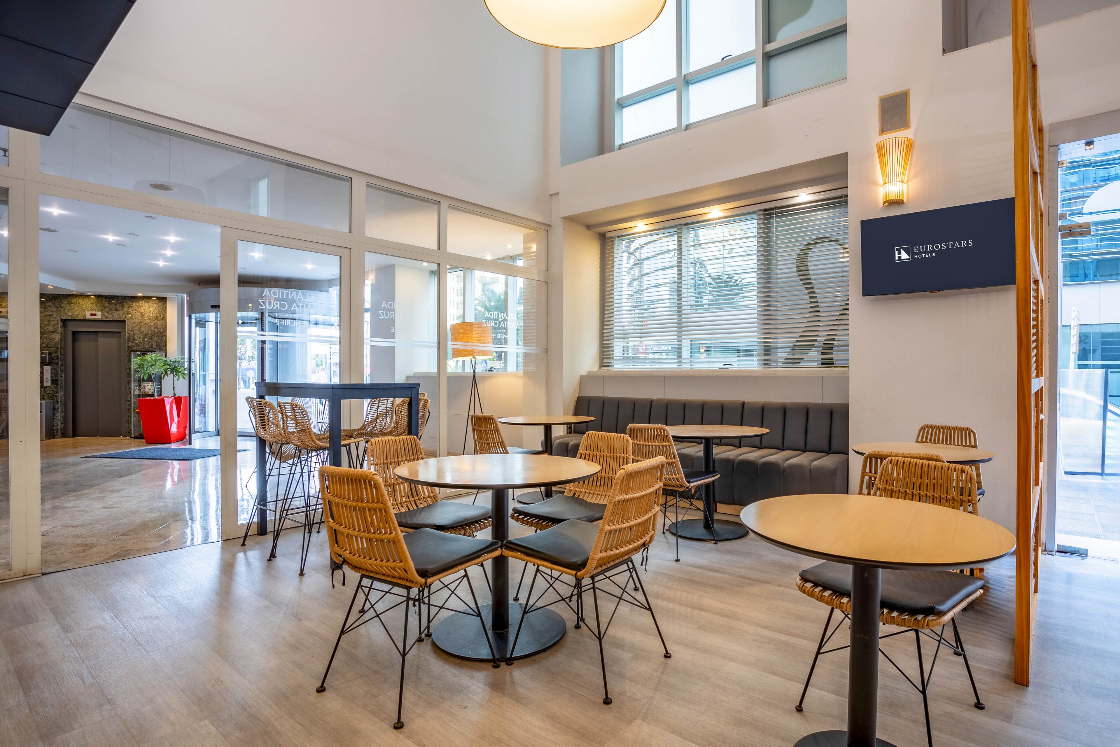 A contemporary cafe space features round wooden tables paired with wicker chairs and a cushioned bench along the wall. The setting is bright and airy, with large windows allowing natural light to fill the room. A digital screen on the wall displays the text 'EUROSTARS,' and the decor includes minimalist lighting and neutral tones. The overall mood is inviting and relaxed, suitable for casual dining or coffee breaks.
