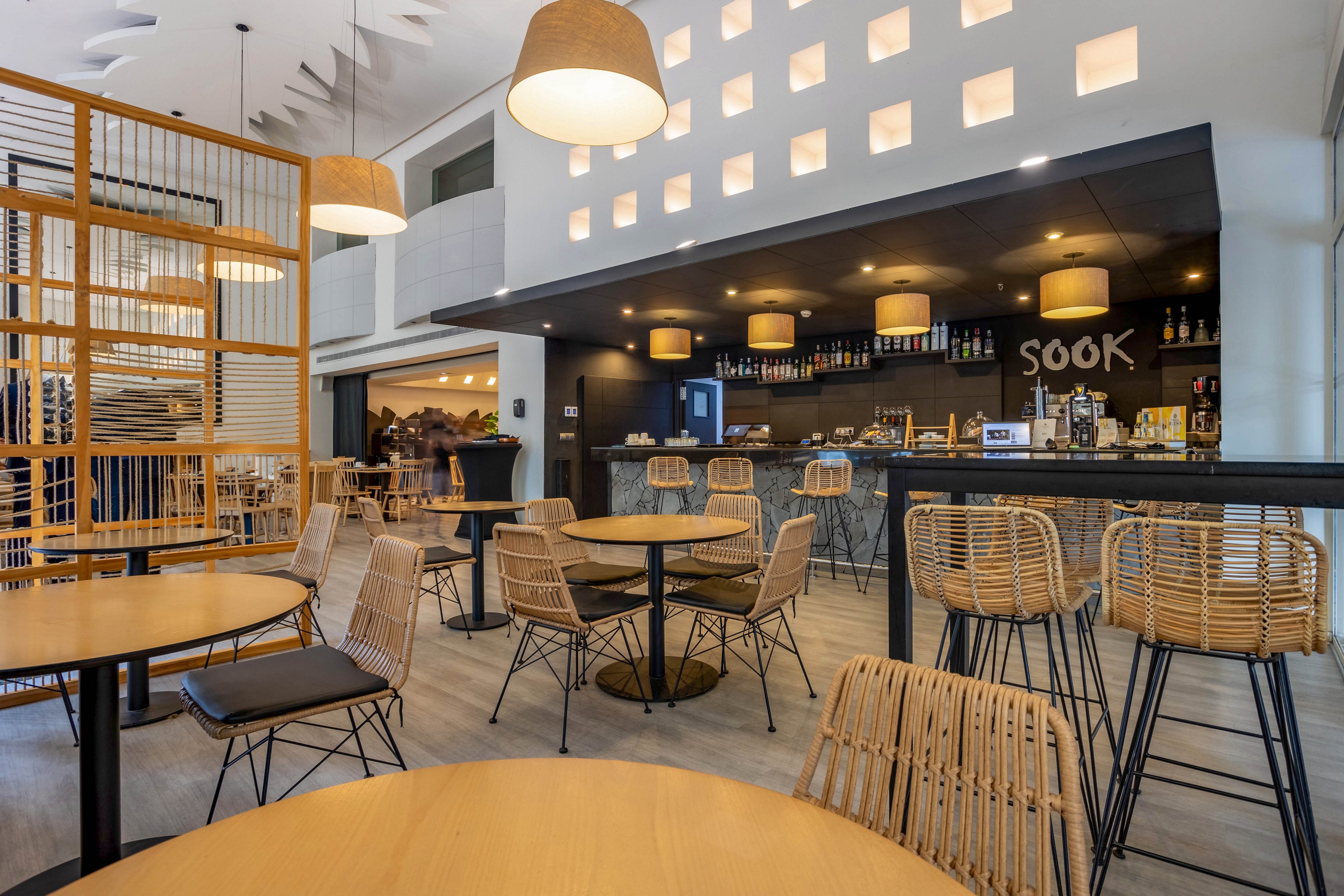The image shows a contemporary café featuring wicker chairs and round wooden tables. The bar area is highlighted by pendant lights and a black counter, with the word 'SOOK' clearly visible on the back wall. The setting is bright and airy, with geometric wall cutouts and a neutral color palette. No people are present, emphasizing the inviting and stylish atmosphere.