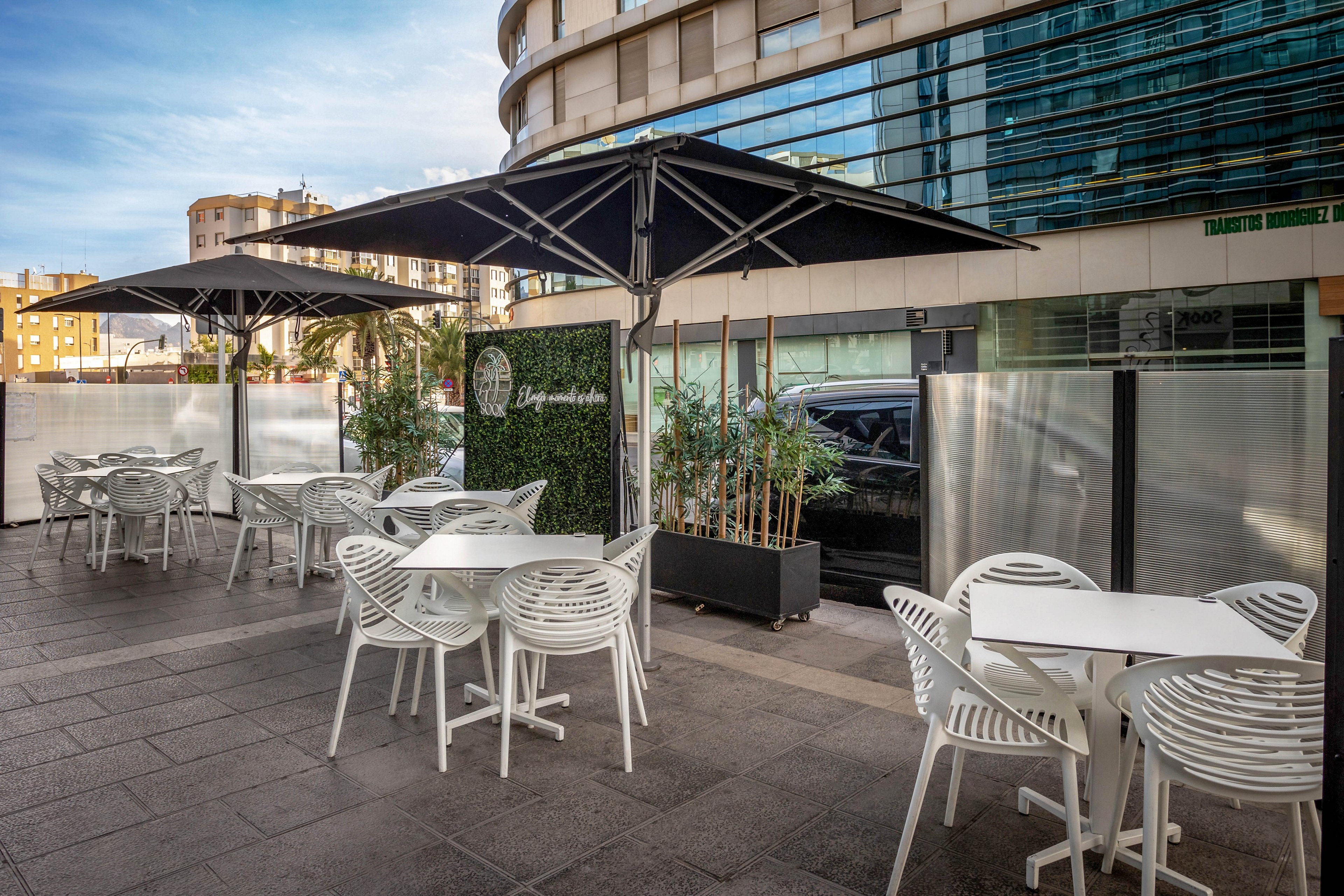 The image shows a contemporary outdoor cafe area featuring white tables and chairs arranged beneath large black umbrellas. The setting is urban, with glass-fronted buildings and potted plants providing a stylish backdrop. The mood is relaxed and inviting, with neutral tones and clean lines dominating the scene. Visible text on the greenery wall includes 'Lounge Bar', adding context to the location.