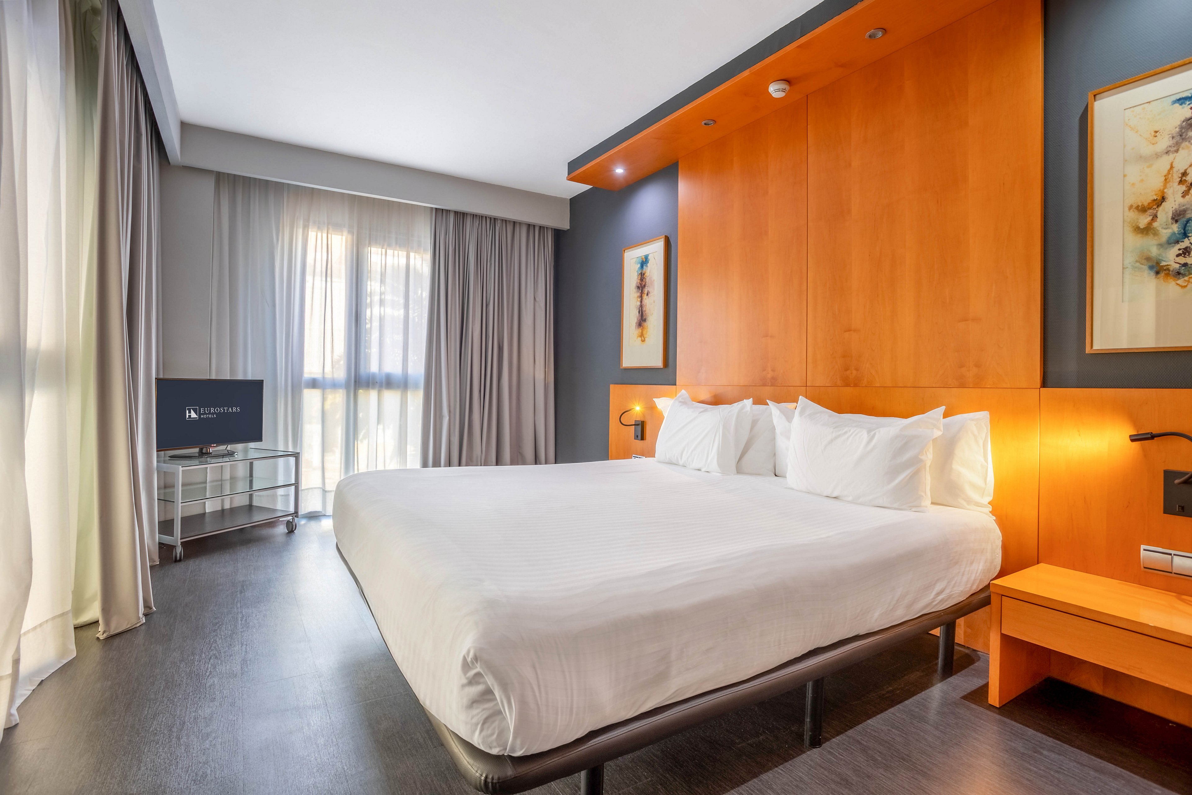 A spacious hotel room features a large bed with white linens and multiple pillows, set against a warm wooden accent wall. Natural light streams in through sheer curtains, illuminating the minimalist decor and contemporary furnishings. A flat-screen television sits on a metal stand, and framed artwork adds a touch of color to the space. The overall mood is calm and inviting, with a neutral palette and clean lines.