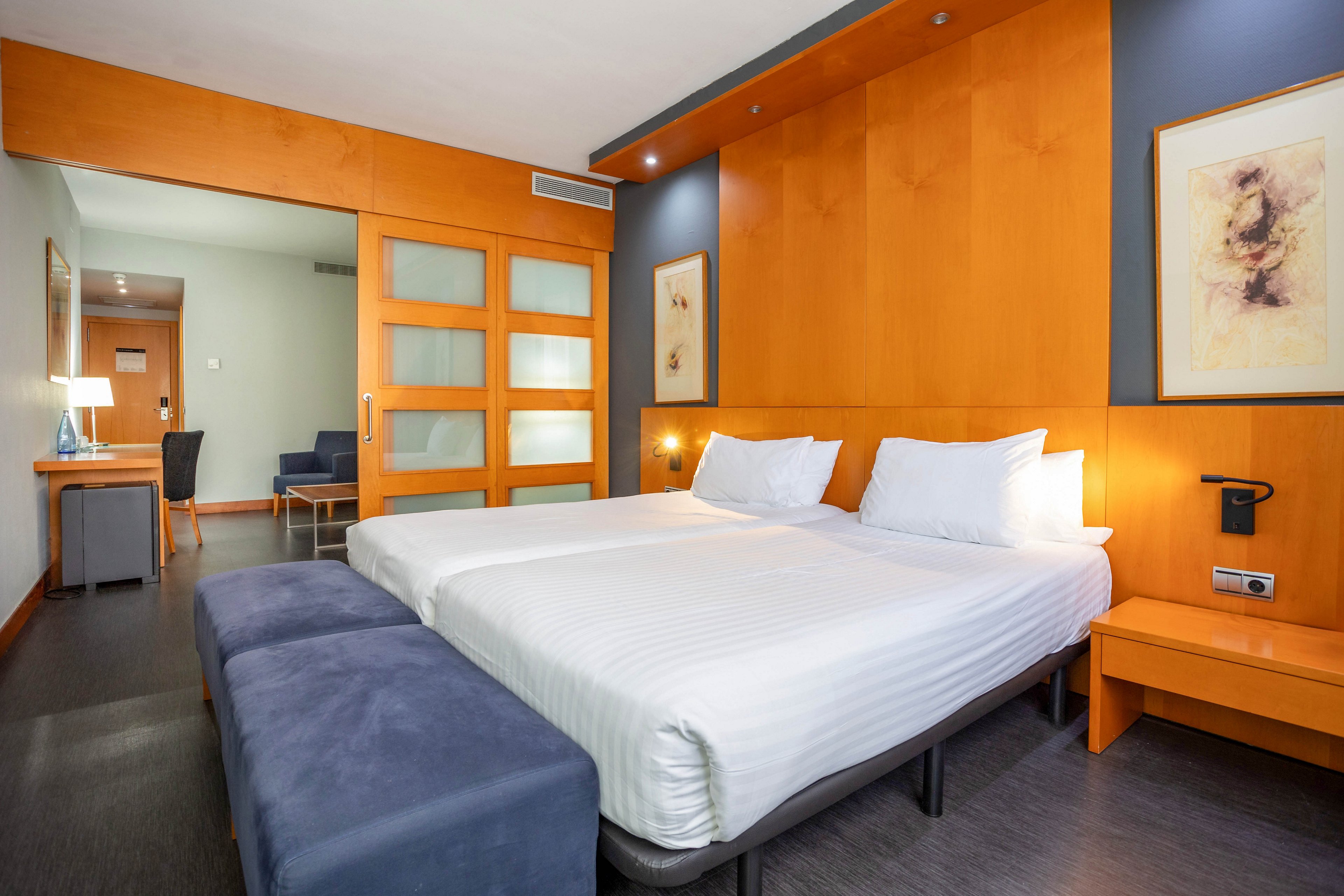 A contemporary hotel room features two neatly made twin beds with white linens, set against a backdrop of warm wooden paneling and blue-gray walls. The room includes a desk, seating area, and framed abstract artwork. Soft lighting and minimalist decor create a calm, inviting atmosphere. No visible text or numeric values are present in the image.