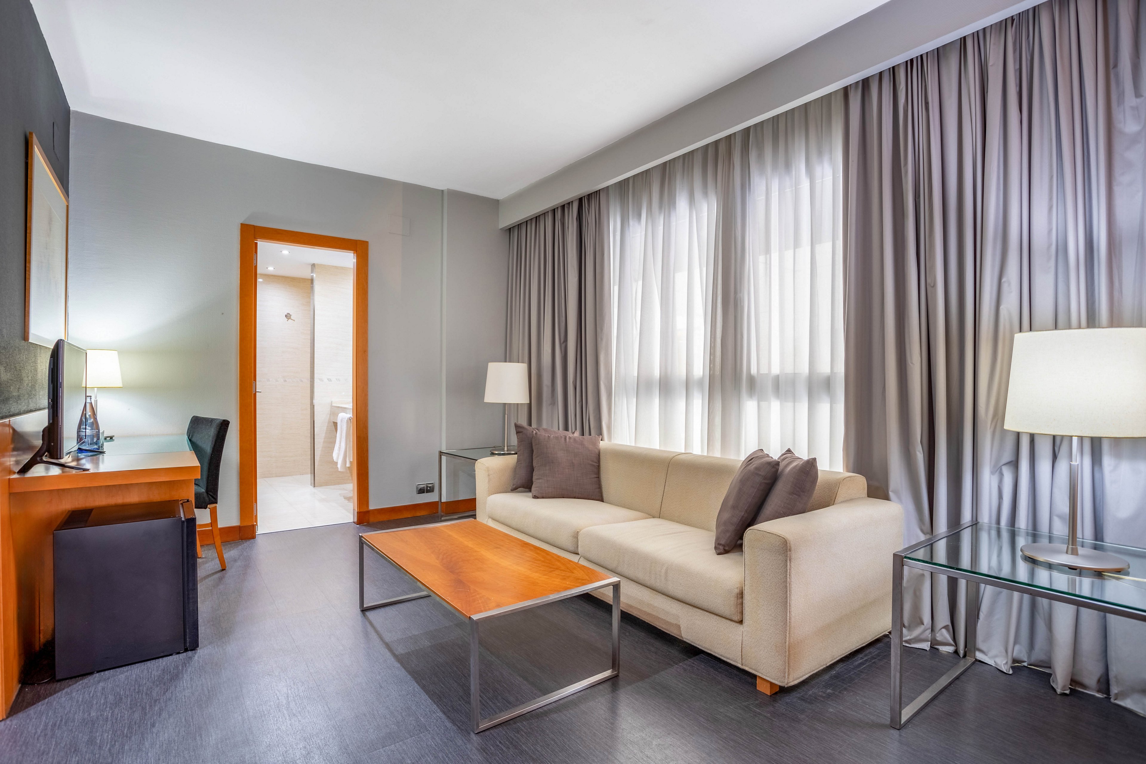 A contemporary hotel suite features a beige sofa, glass tables, and a wooden coffee table. The room includes a workspace with a desk and chair, and large windows with gray curtains allow natural light to fill the space. Soft lighting from table lamps creates a calm and inviting atmosphere, and an open doorway leads to a bathroom area.