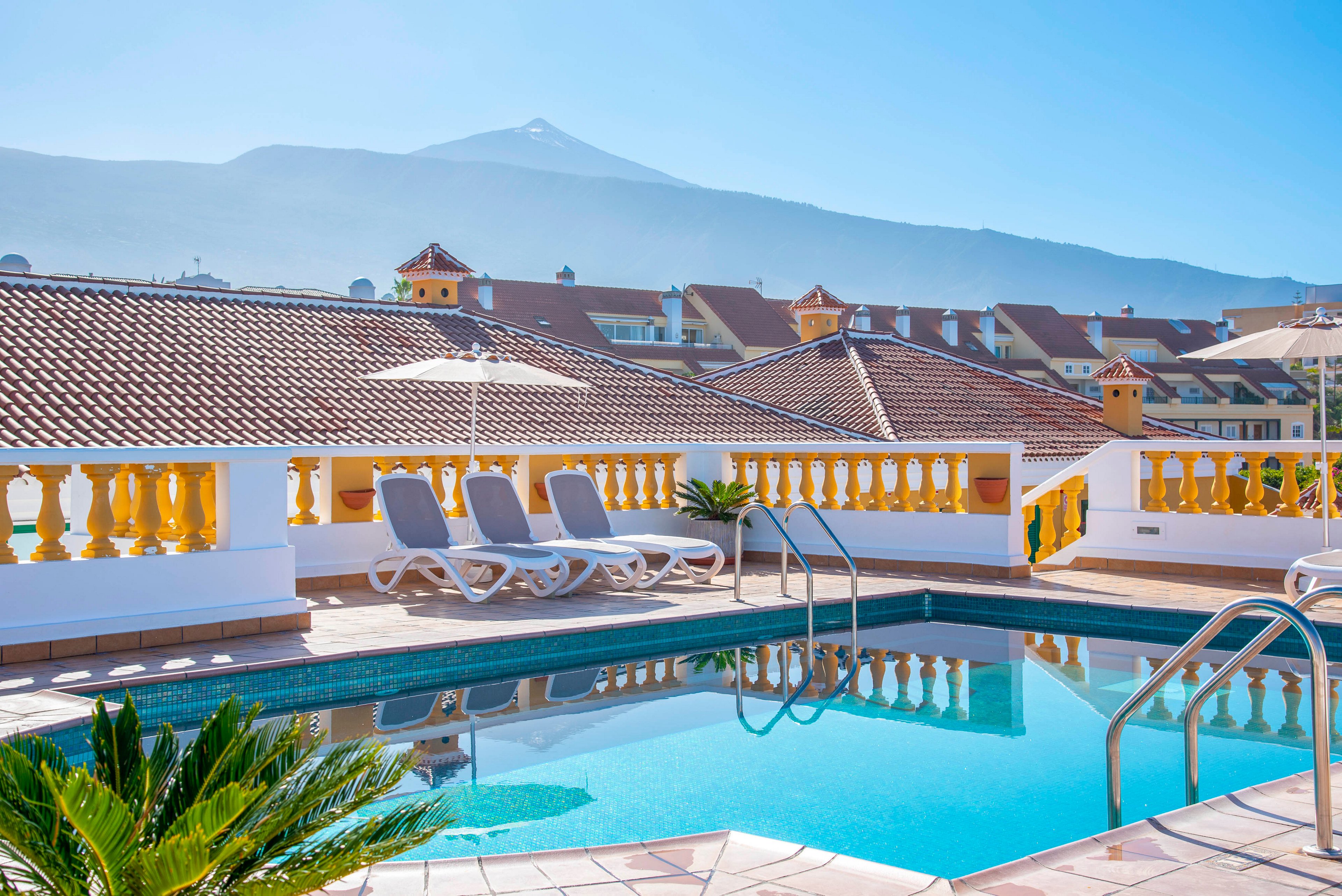 A clear blue swimming pool sits on a sunny rooftop terrace with several empty lounge chairs. Decorative yellow railings and tiled roofs of nearby buildings frame the scene. In the background, a tall mountain rises under a bright blue sky. The calm water and clear weather create a relaxing vacation atmosphere.