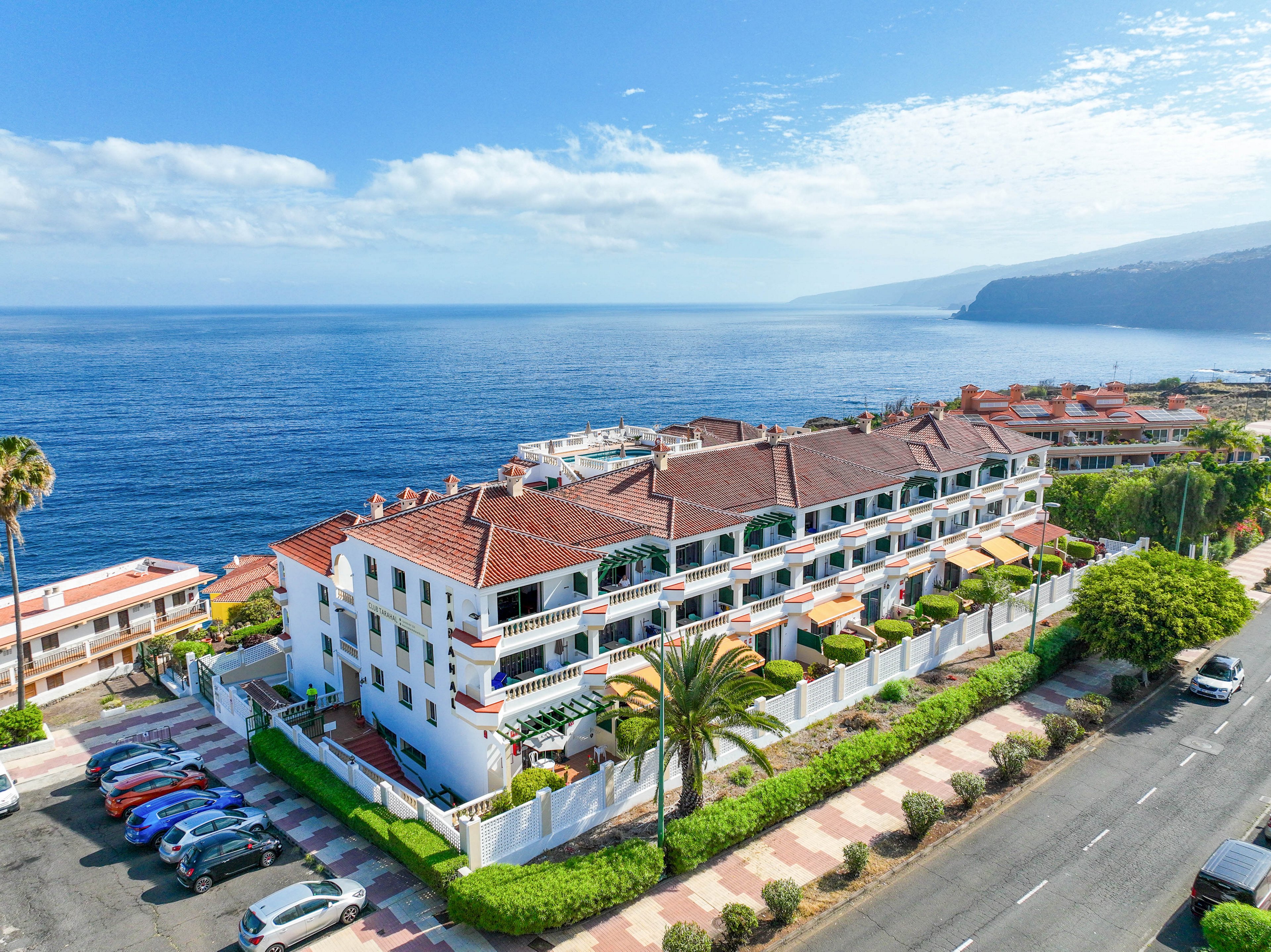 A large white apartment complex with red-tiled roofs sits along a coastal road overlooking a calm blue sea. The building features multiple balconies and terraces facing the ocean, surrounded by palm trees and manicured greenery. Parked cars line the street in front, while additional seaside buildings extend into the distance. Clear skies and scattered clouds create a bright, inviting holiday atmosphere.