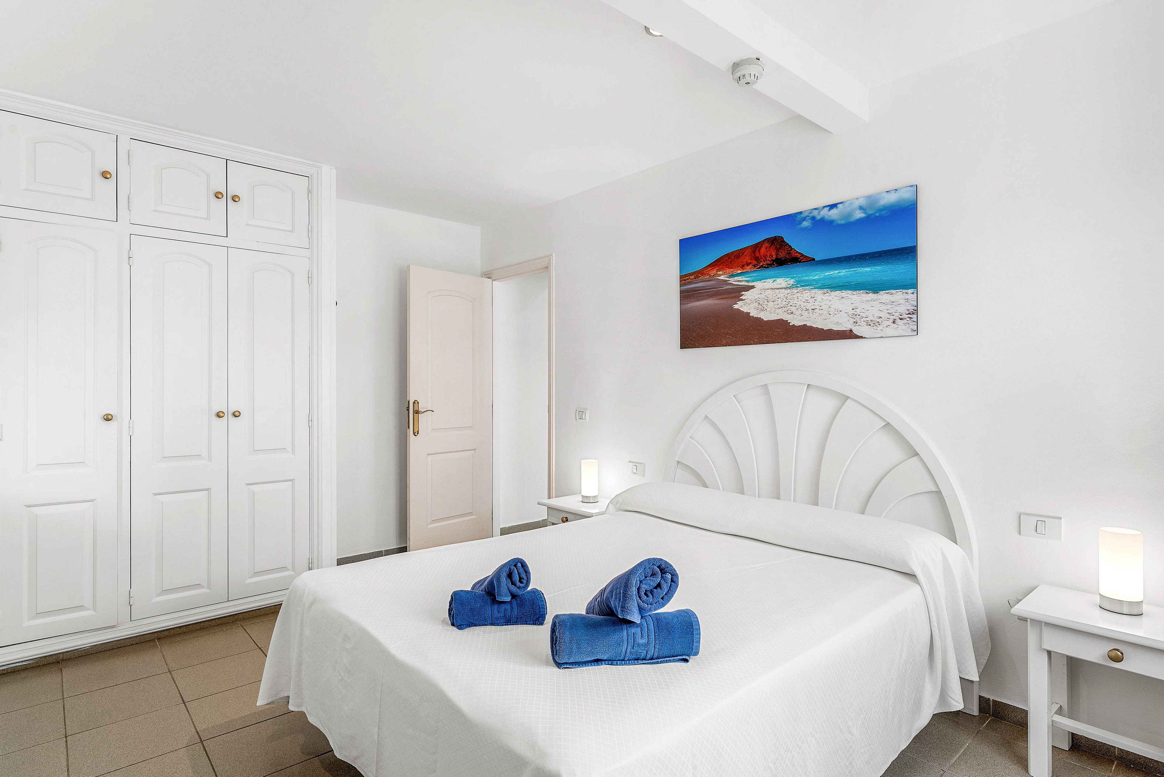 A clean white bedroom features a neatly made double bed with a curved headboard. Two rolled blue towels are placed on the bed, adding a pop of color to the neutral space. Built-in white wardrobes line one wall, and bedside tables with small lamps flank the bed. A vivid beach and mountain photograph hangs above the headboard, enhancing the calm, coastal-inspired atmosphere.