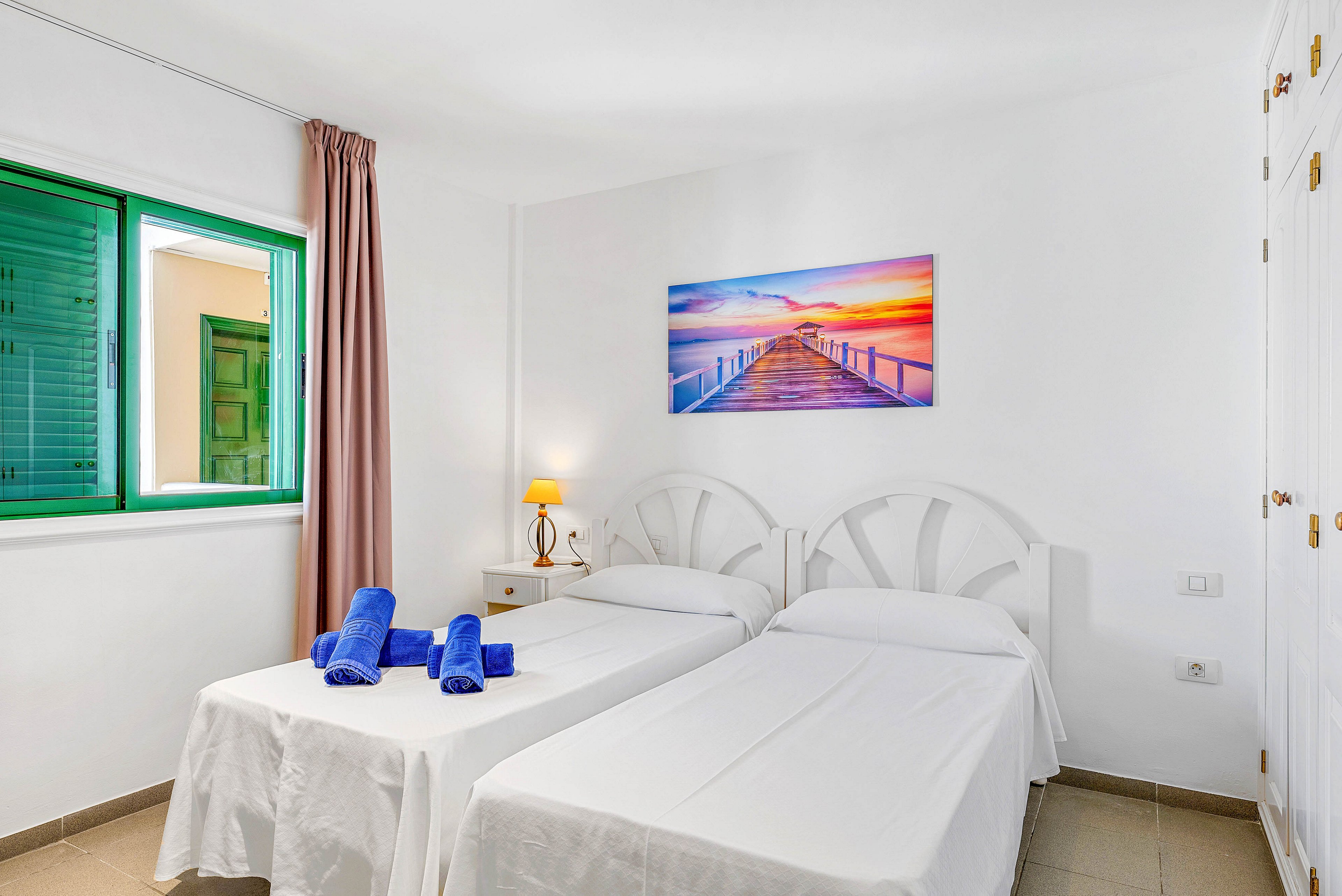 A clean, white twin bedroom features two single beds with simple white linens. Rolled blue towels are neatly placed on one of the beds, adding a pop of color. A small bedside table with a lit lamp sits between the beds, beneath a colorful sunset pier photograph on the wall. Natural light enters through a green-framed window with a mauve curtain, creating a fresh and inviting atmosphere.