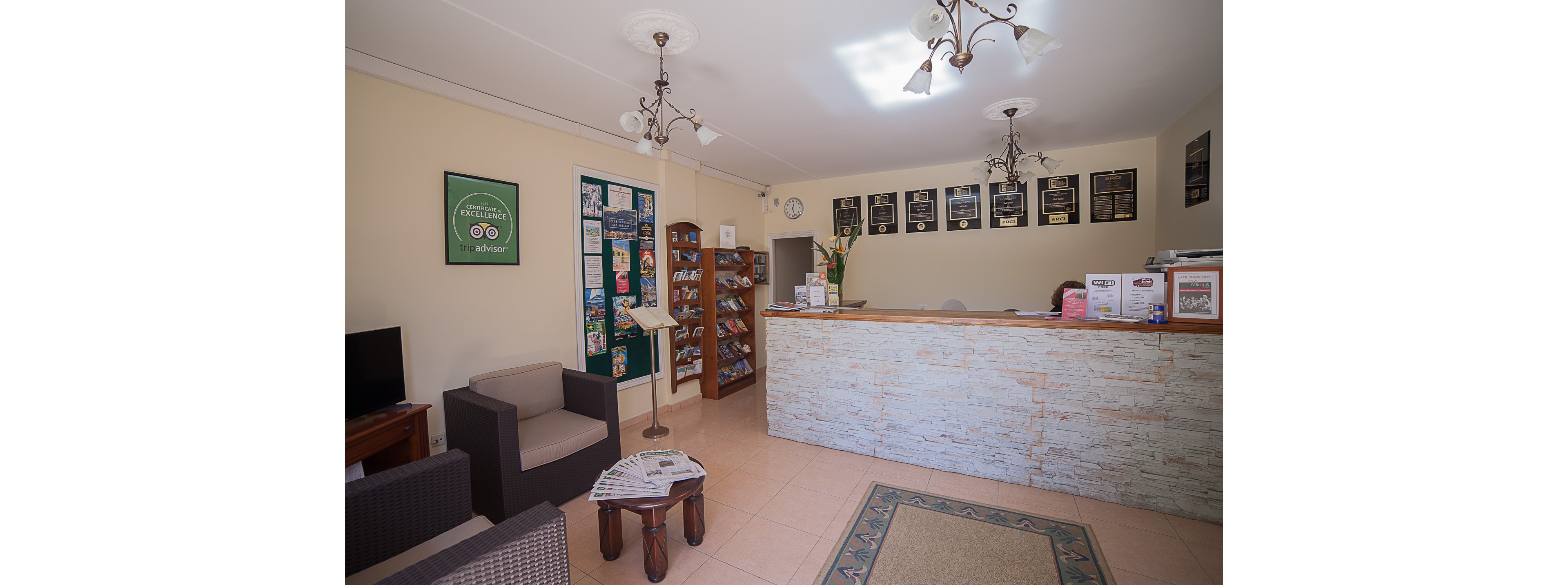 A small hotel reception area features a stone-front desk with brochures and paperwork neatly arranged on top. Behind the counter, framed certificates and a wall clock decorate the light-colored wall. To the left, a seating area with armchairs, a small table, and a television offers a comfortable waiting space. A brochure stand and bulletin board filled with travel information add to the welcoming, informative atmosphere.