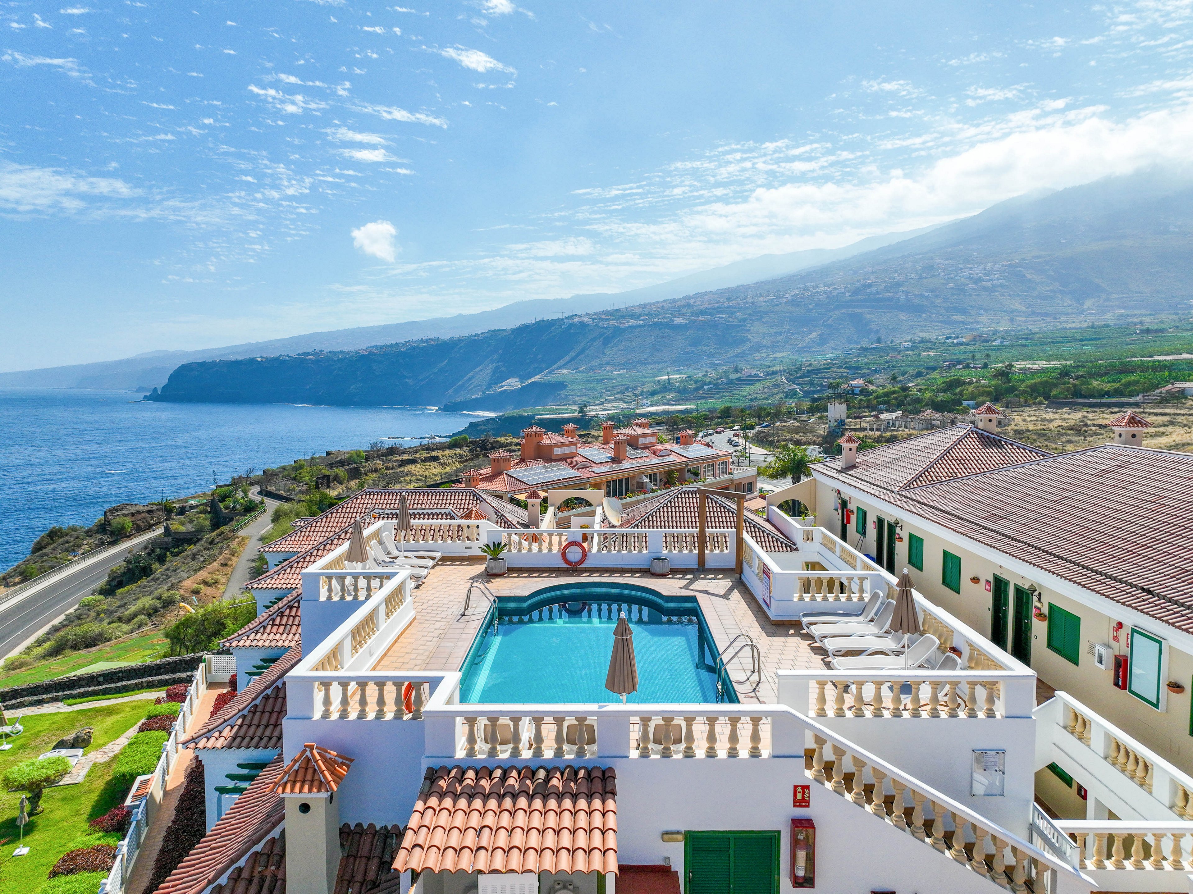 A hilltop hotel features a central outdoor swimming pool surrounded by sun loungers and white balustrades. The building’s terracotta roofs and green shutters contrast with the bright blue water of the pool. In the background, a winding coastal road leads toward dramatic sea cliffs and a calm ocean. Rolling hills and scattered houses stretch into the distance under a lightly clouded sky, creating a serene vacation atmosphere.