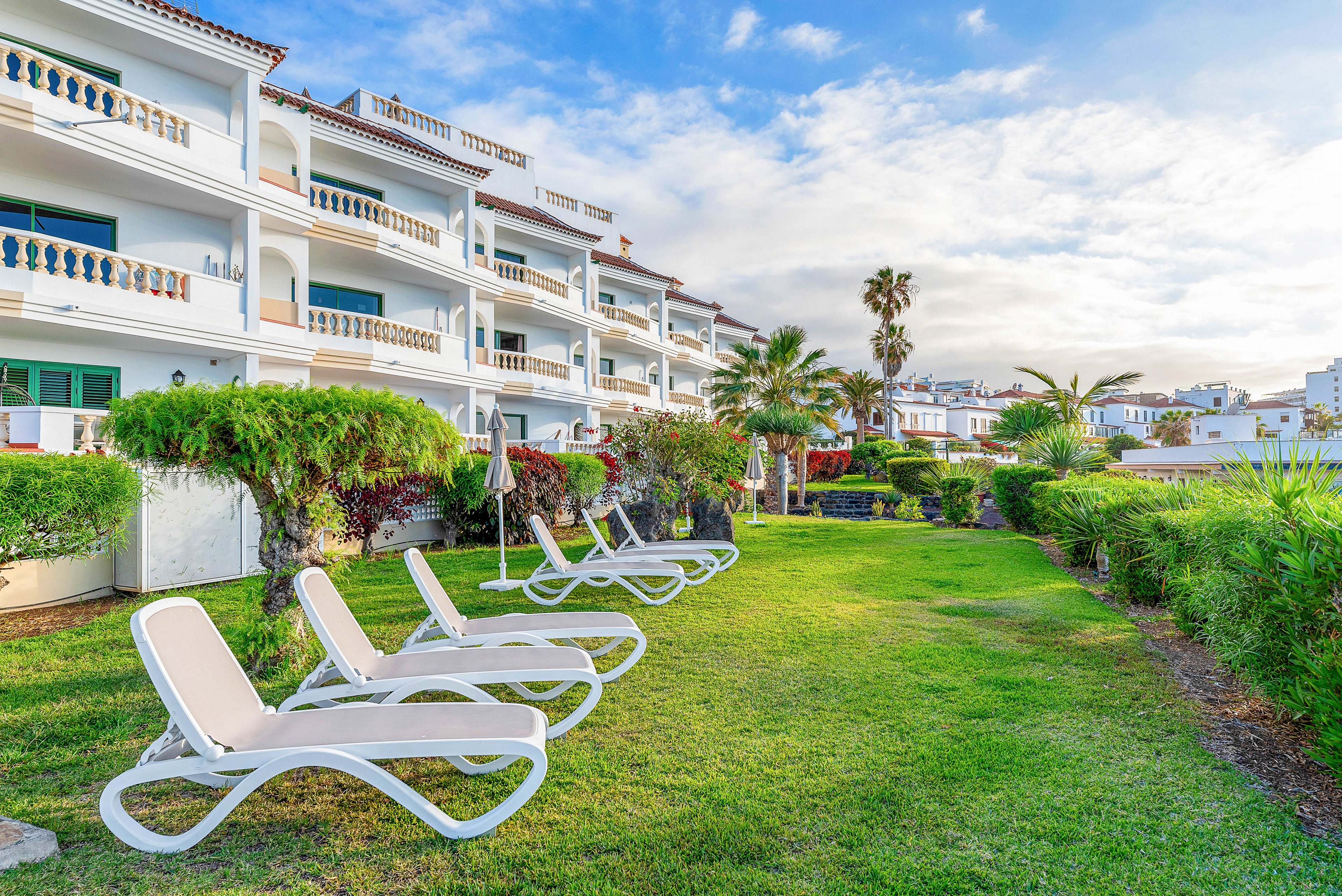 A row of white sun loungers is arranged on a neatly manicured lawn beside a multi-story Mediterranean-style building. The resort features white facades, balconies with railings, and tiled roofs. Lush green shrubs, palm trees, and other tropical plants line the garden, creating a relaxing vacation atmosphere. The sky is partly cloudy with soft sunlight illuminating the scene, suggesting a warm coastal destination.