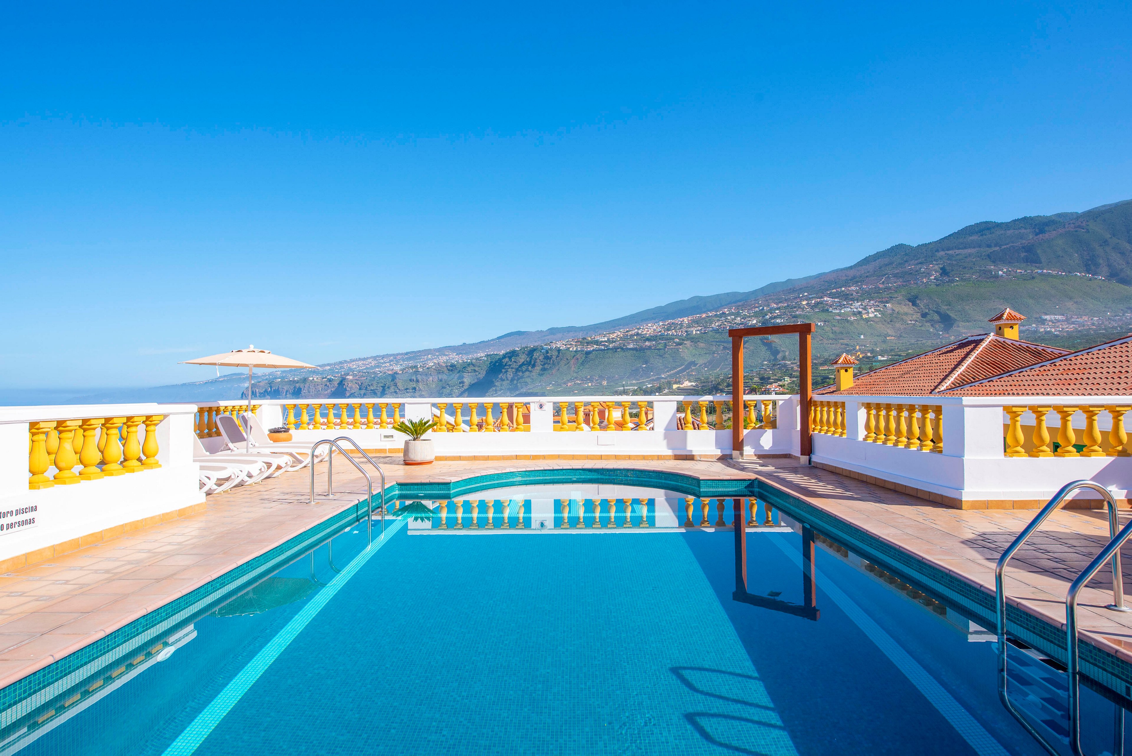 An outdoor swimming pool sits on a sunny rooftop terrace with clear blue water. White and yellow balustrades frame the deck, with lounge chairs and a sun umbrella arranged for relaxation. In the distance, green mountains and scattered houses rise under a bright blue sky. The scene suggests a tranquil vacation setting with panoramic coastal views.