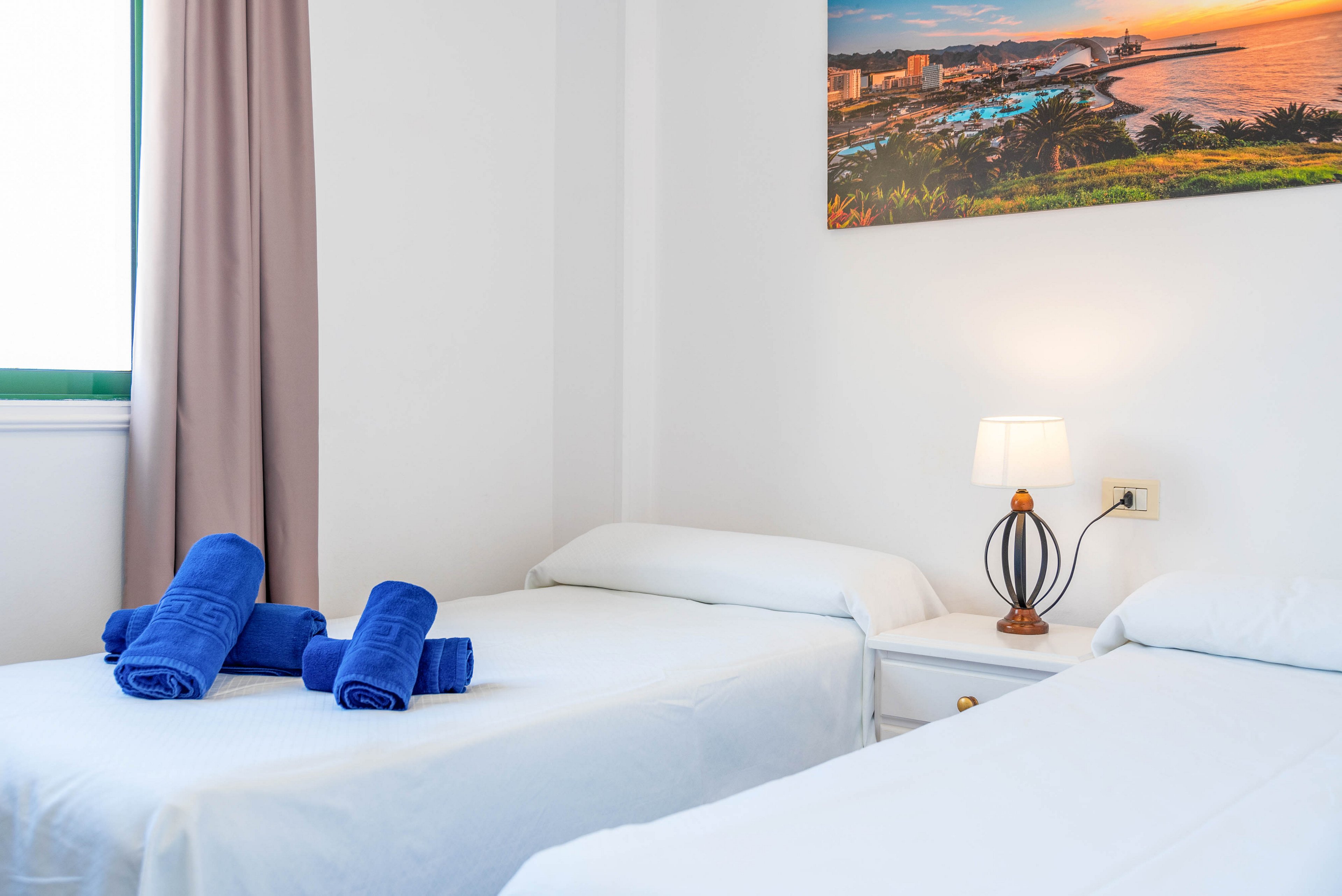 A clean, minimal twin bedroom features two single beds with crisp white linens. Rolled blue towels are neatly placed on one of the beds, adding a pop of color. A small bedside table with a decorative lamp sits between the beds, casting a warm light. A large framed photograph of a coastal cityscape hangs on the wall, enhancing the modern, inviting atmosphere.